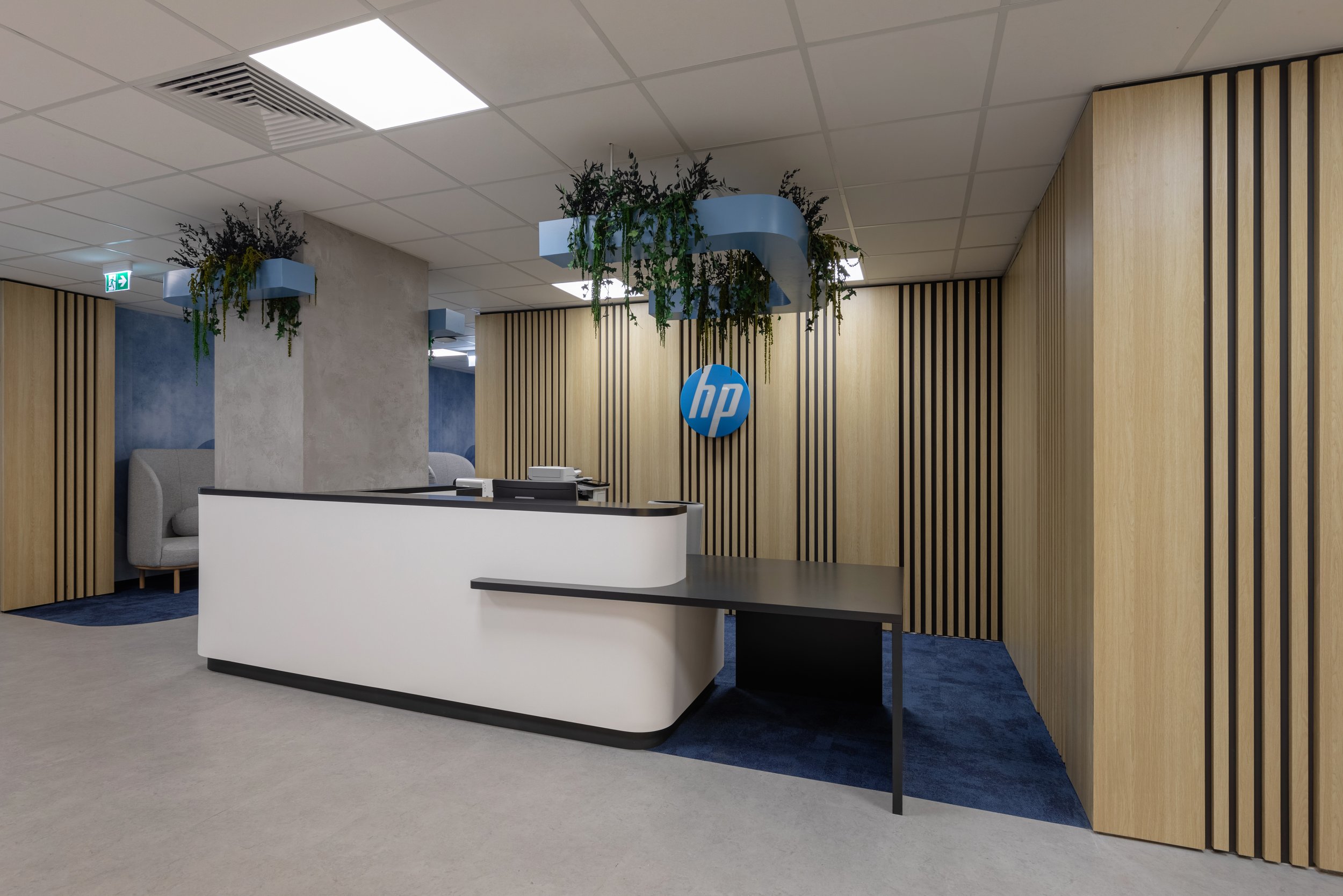 Office reception area featuring a white curved reception desk, HP logo on a wooden-paneled wall, and blue ceiling decor with hanging plants.