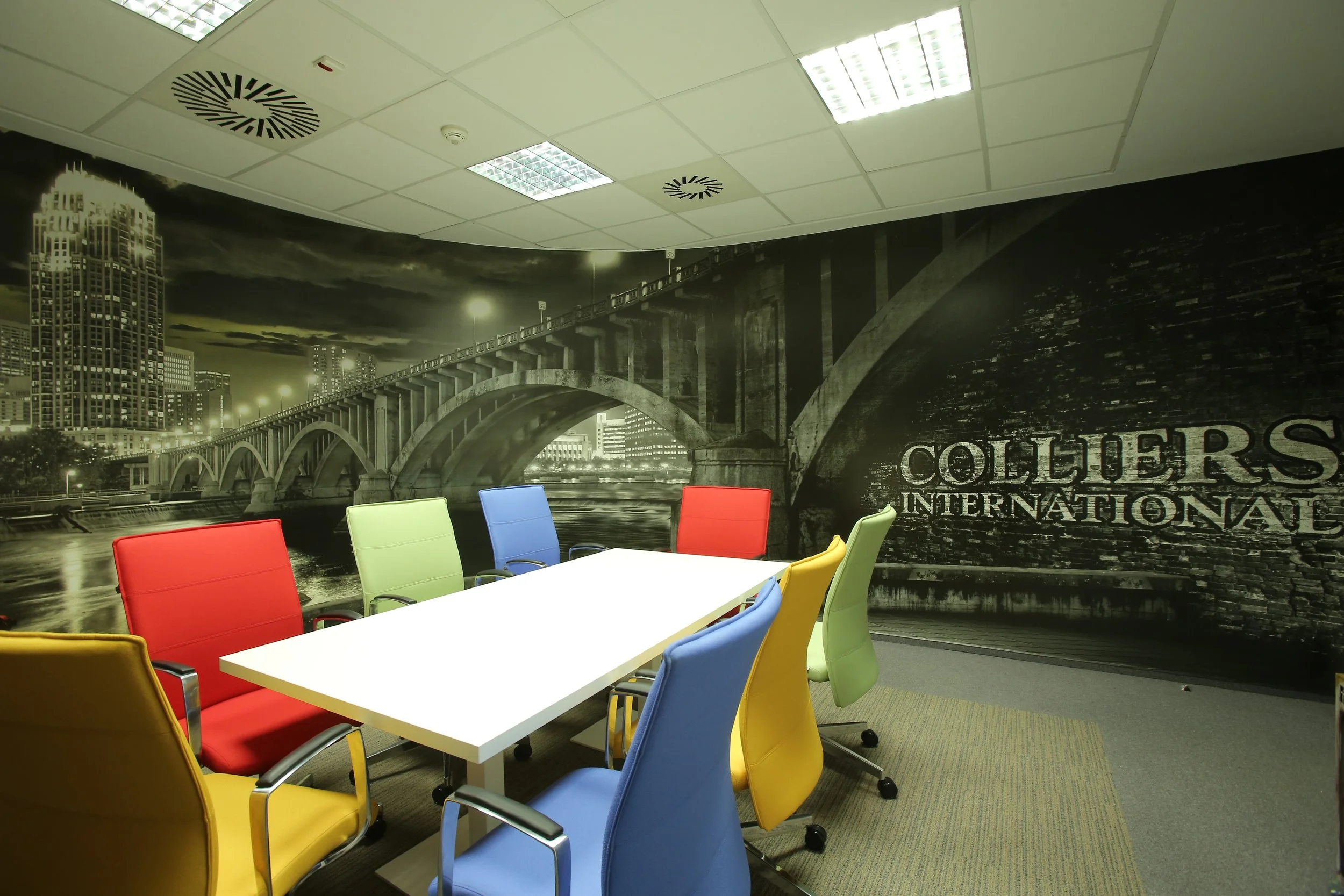 Conference room with colorful chairs around a white table, and a black and white cityscape mural on the wall reading 'COLLIERS INTERNATIONAL'.