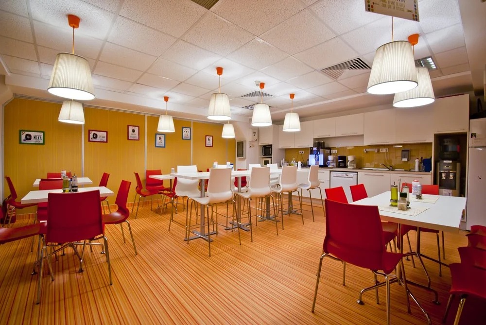 A bright breakfast room with white tables, red and white chairs, yellow walls, and hanging white lamps.