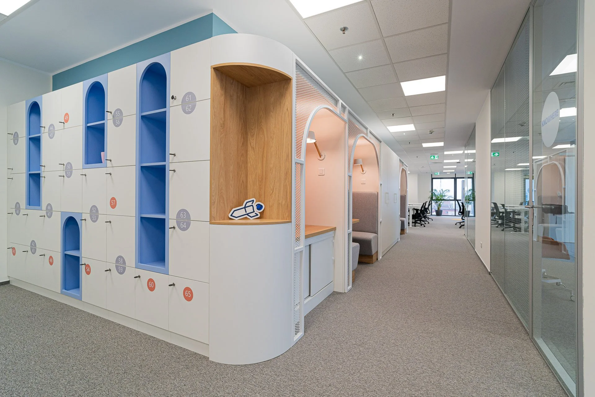 Modern office space with labeled lockers on the left, private seating booths along the corridor, and glass walls on the right, with chairs and plants in the background.