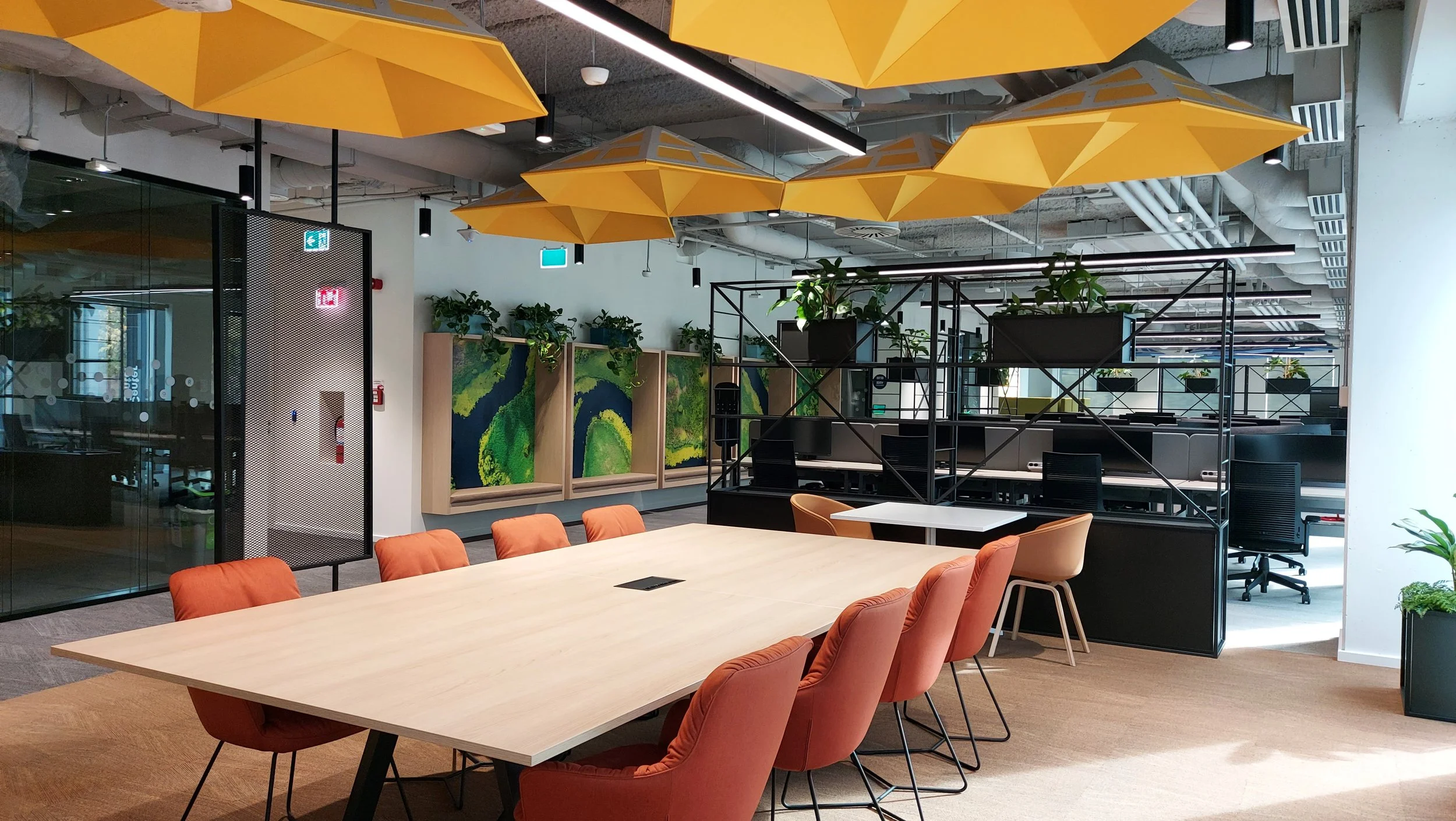 Modern office conference room with orange chairs around a wooden table, yellow geometric ceiling panels, greenery, and open workspace area in the background.
