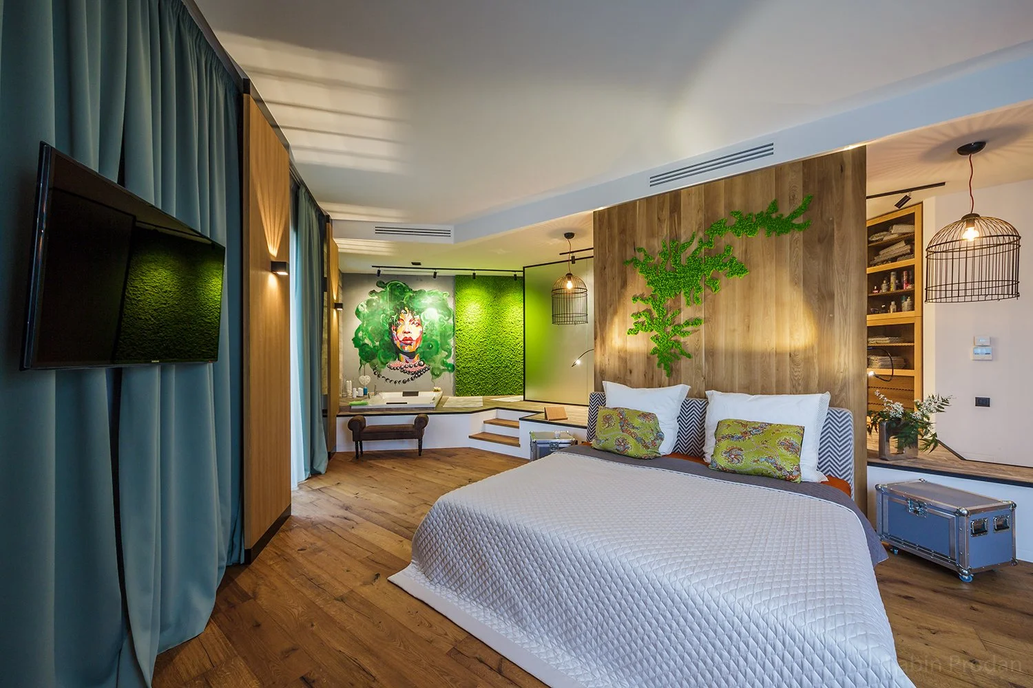 Modern bedroom with wooden floor, white bed, colorful pillows, green wall art, and decorative lighting fixtures.