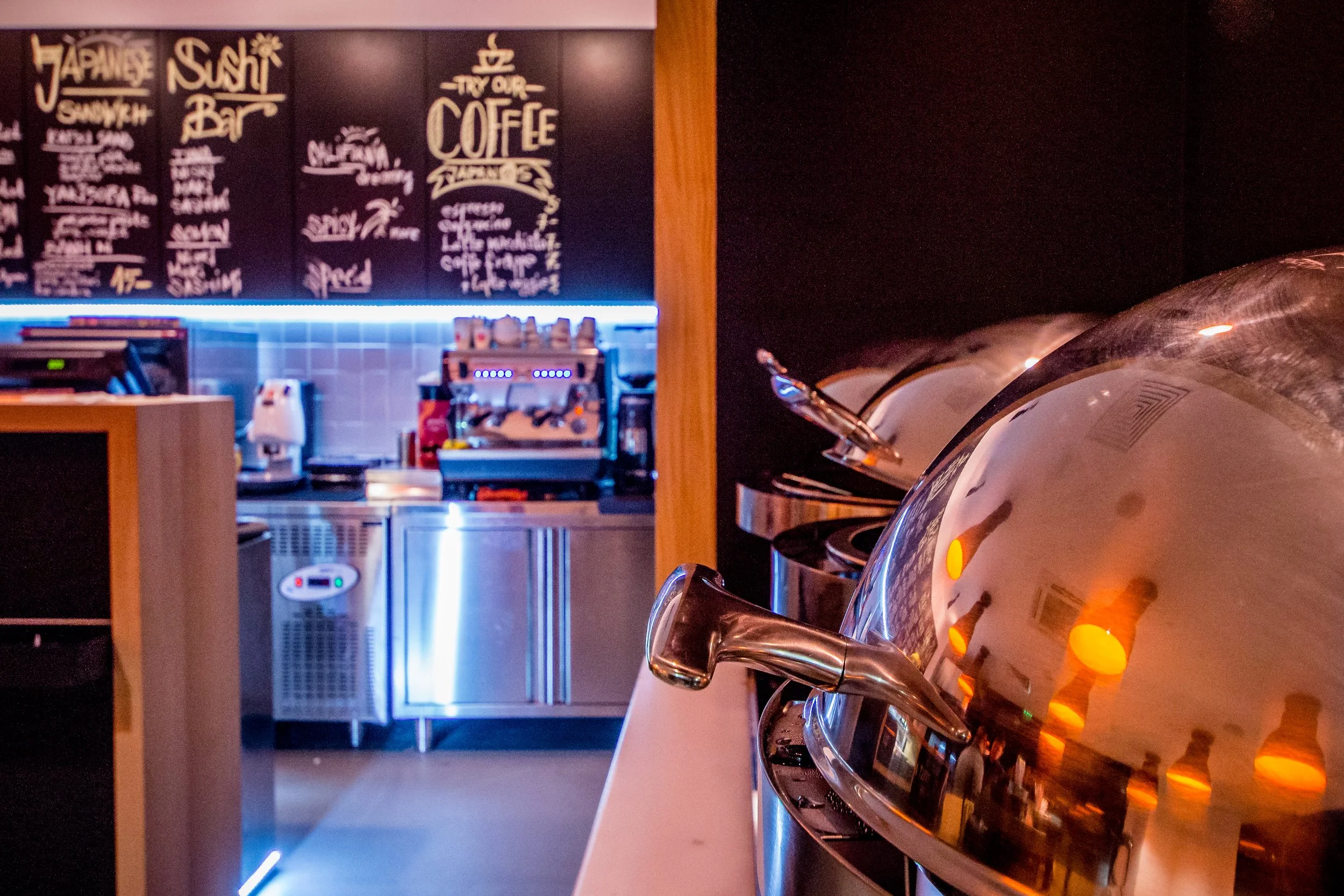 Close-up of a stainless steel coffee urn in a coffee shop, with a dark wall and menu board in the background.