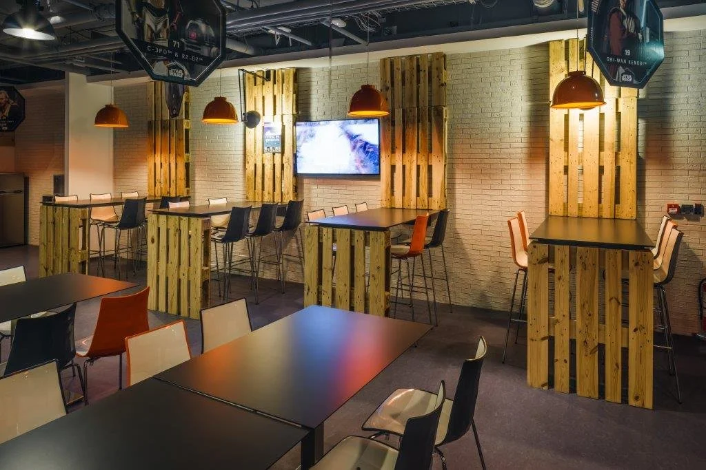 Modern cafe or restaurant interior with black tables, multicolored chairs, wooden decorative panels on the wall, hanging orange lamps, and a TV screen on the wall.