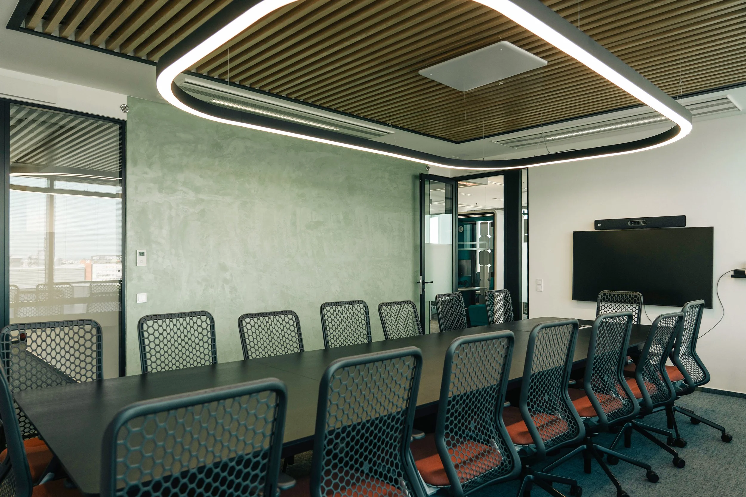 Modern conference room with a long black table, black mesh chairs with orange seats, a large wall-mounted screen with a camera above, and a ceiling with an oval-shaped light fixture in an office with glass walls and city view.