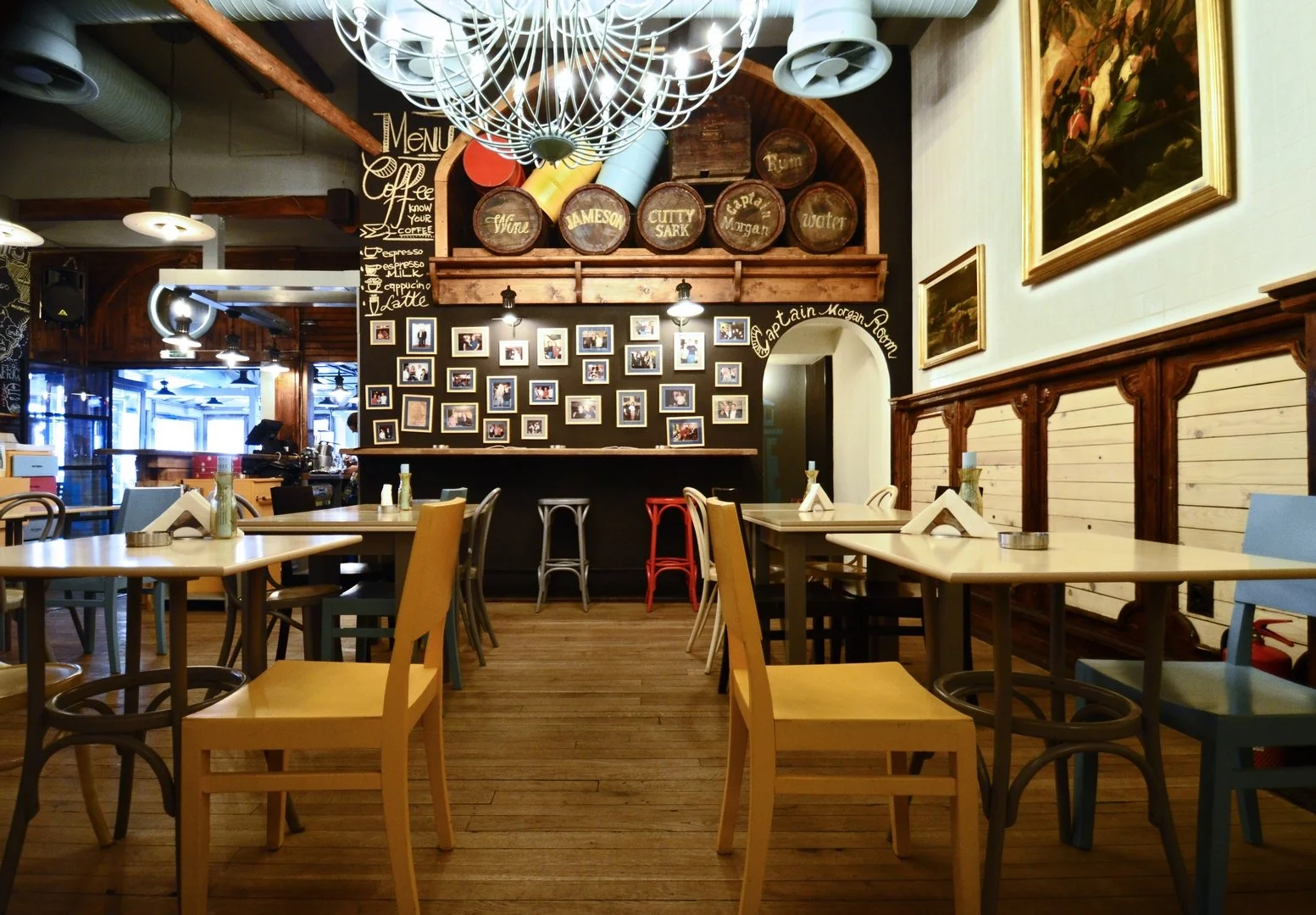 Interior of a cafe or restaurant with wooden floors, various colorful chairs, tables set with tableware, and a black chalkboard wall displaying photos and menu items. Decorative barrels are mounted on a wooden shelf above. Artwork hangs on the wall to the right.