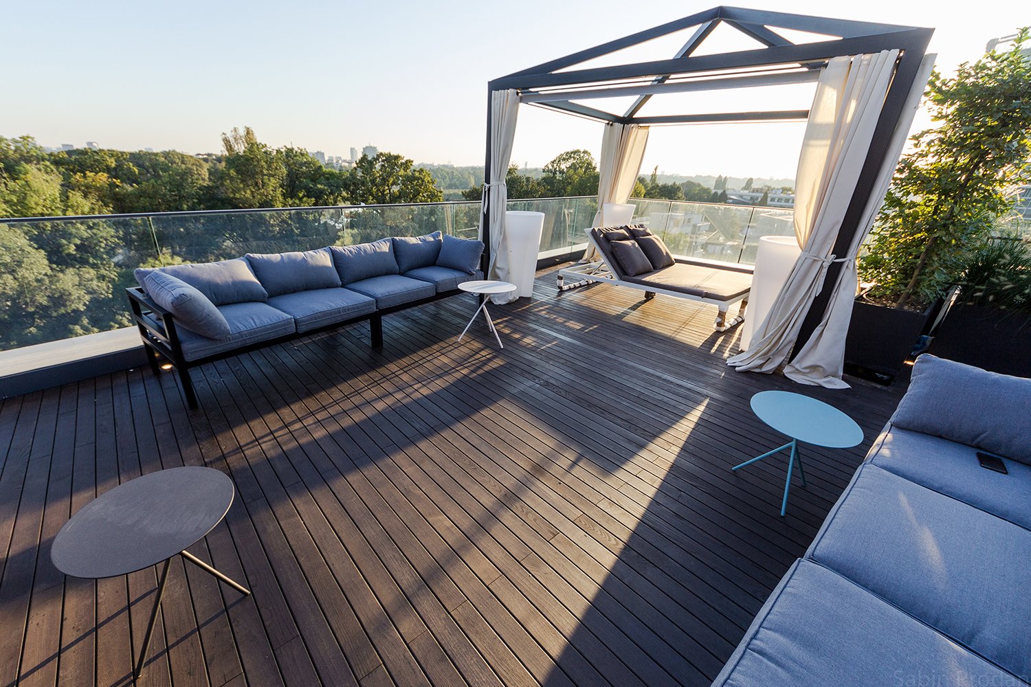 Modern rooftop terrace with outdoor furniture including a large cushioned sofa, a daybed with pillows inside a canopy, and small round tables on wooden decking, overlooking a city skyline with trees and buildings in the distance.