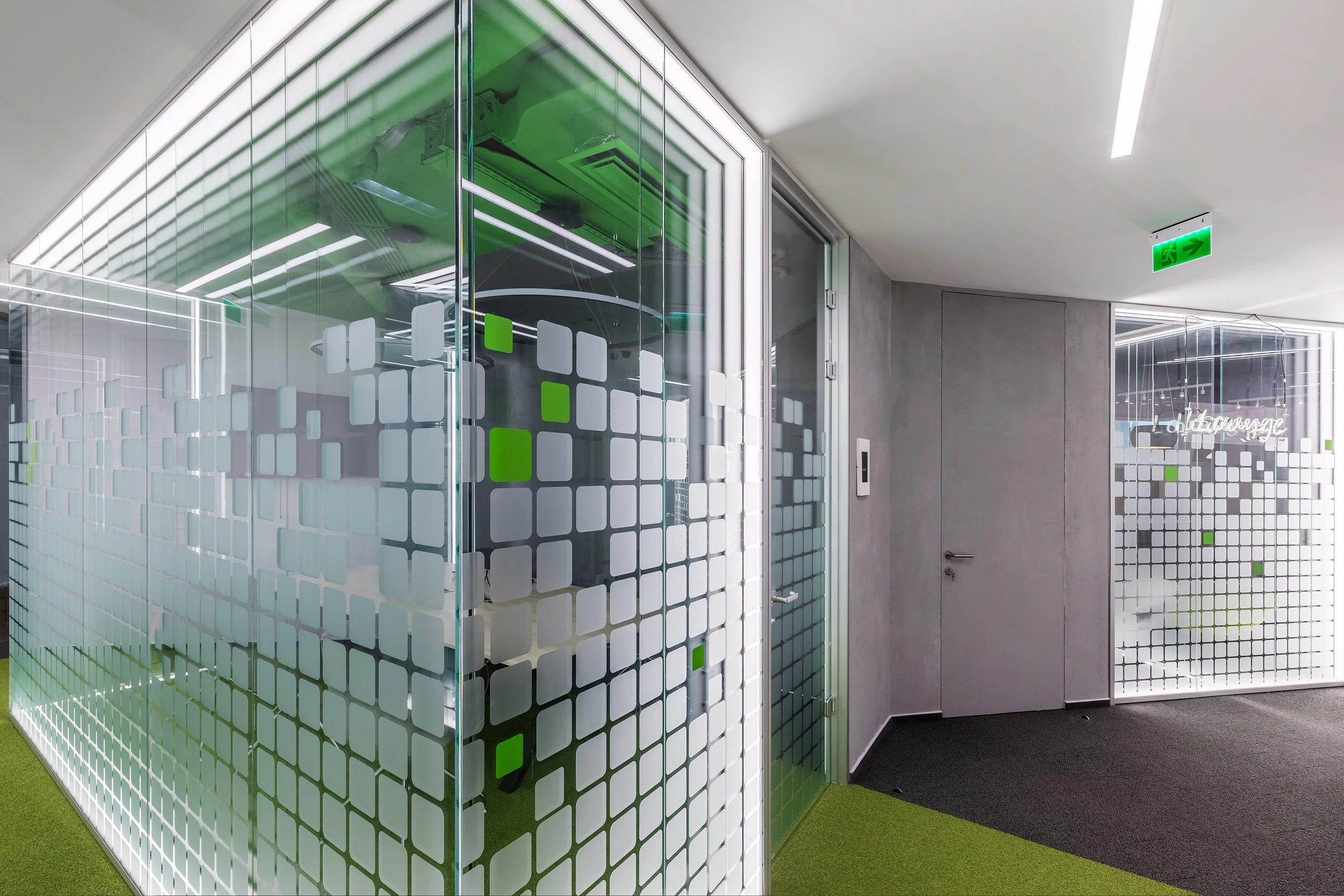 Office hallway with glass-partitioned rooms, one with frosted glass featuring white and green square patterns, green carpet, gray walls, and an illuminated green exit sign.