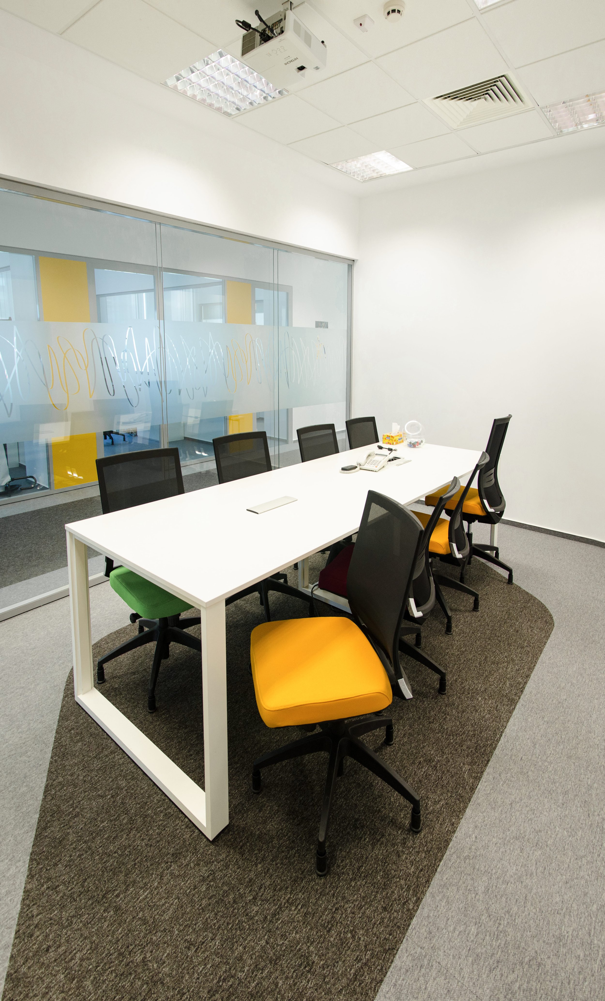 Empty conference room with a white table, six black chairs with yellow, green, and maroon cushions, and a glass wall with abstract yellow and white patterns.