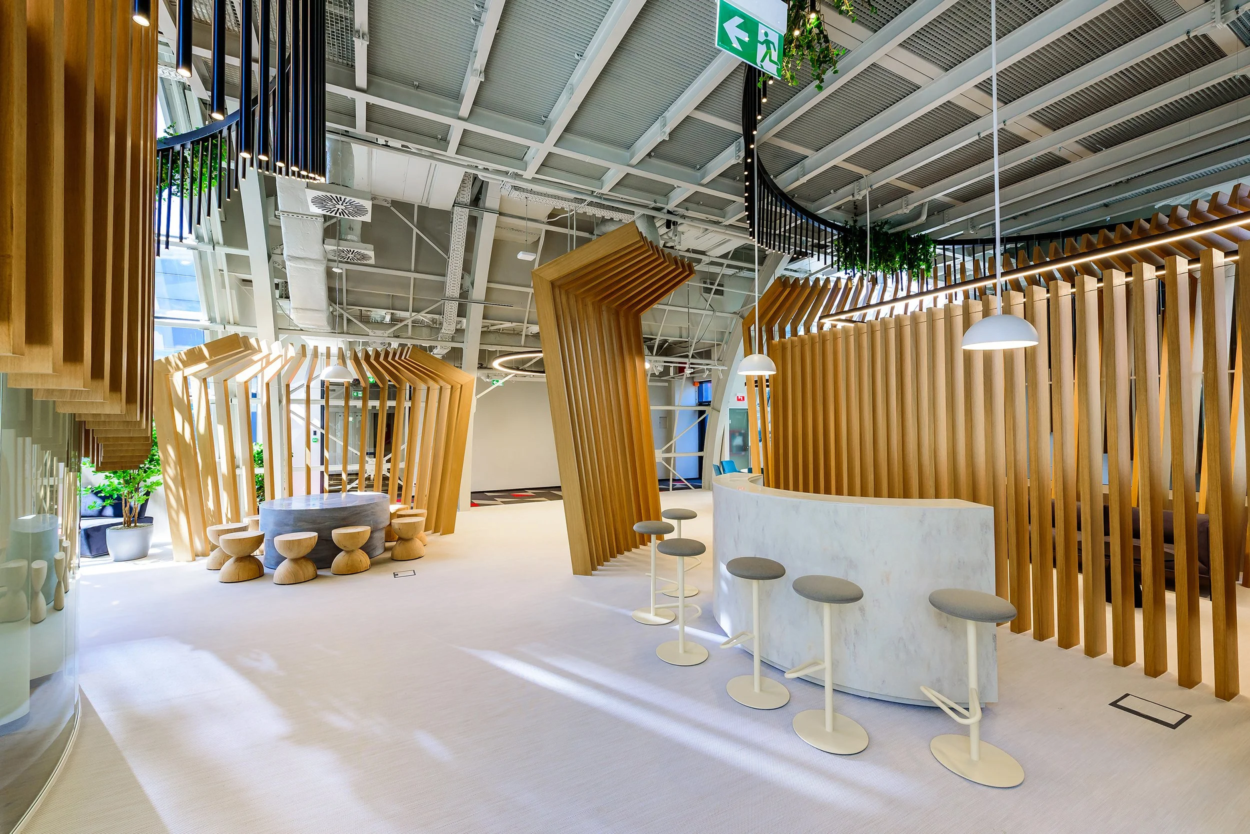 Modern office interior with wooden slatted partitions, white bar counter with stools, and seating area with stools around a table. Overhead lighting and ceiling fixtures are visible, with some greenery hanging from the ceiling.
