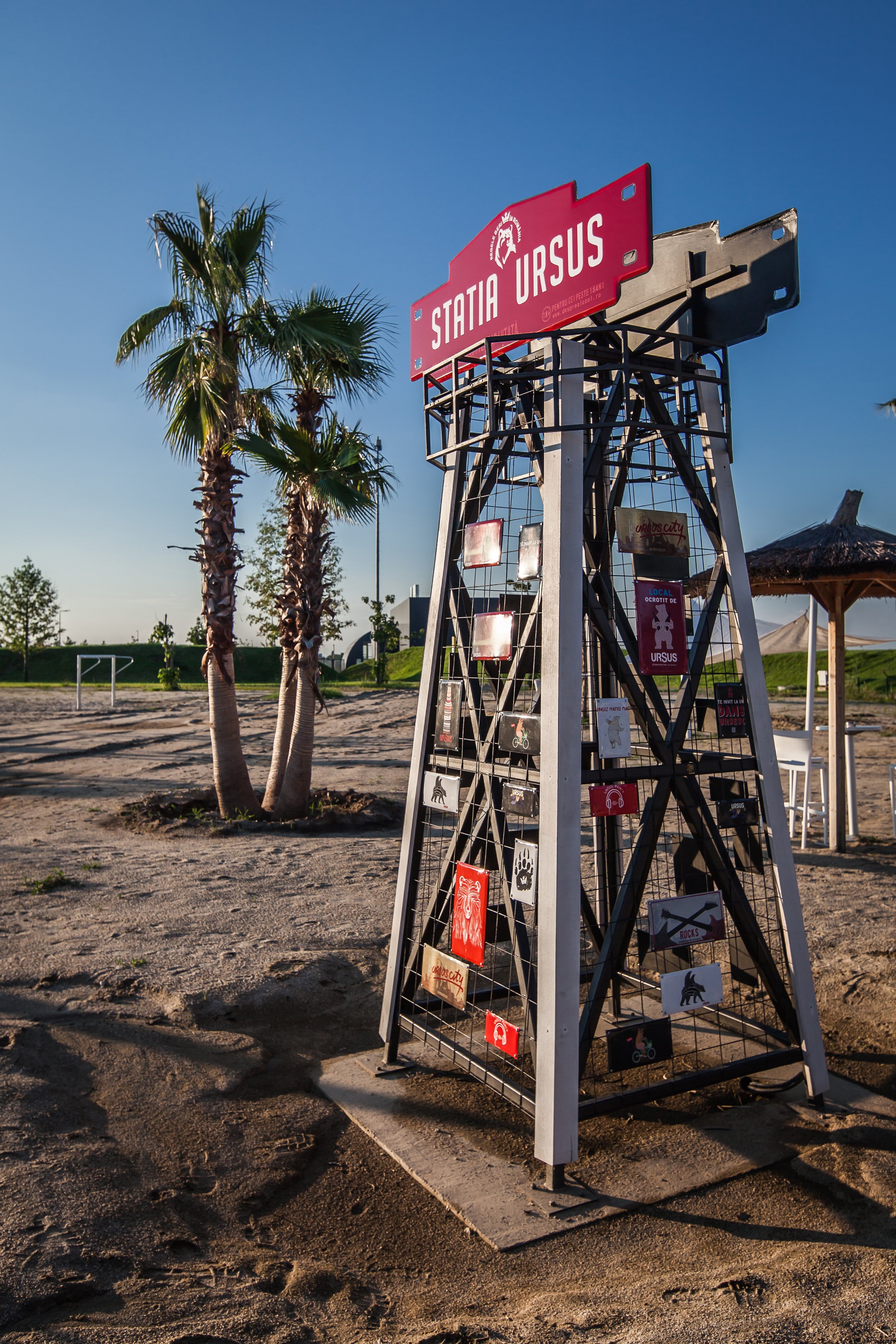 A metal stand with signs stands on sandy ground in a park with palm trees and a thatched umbrella in the background during sunset.