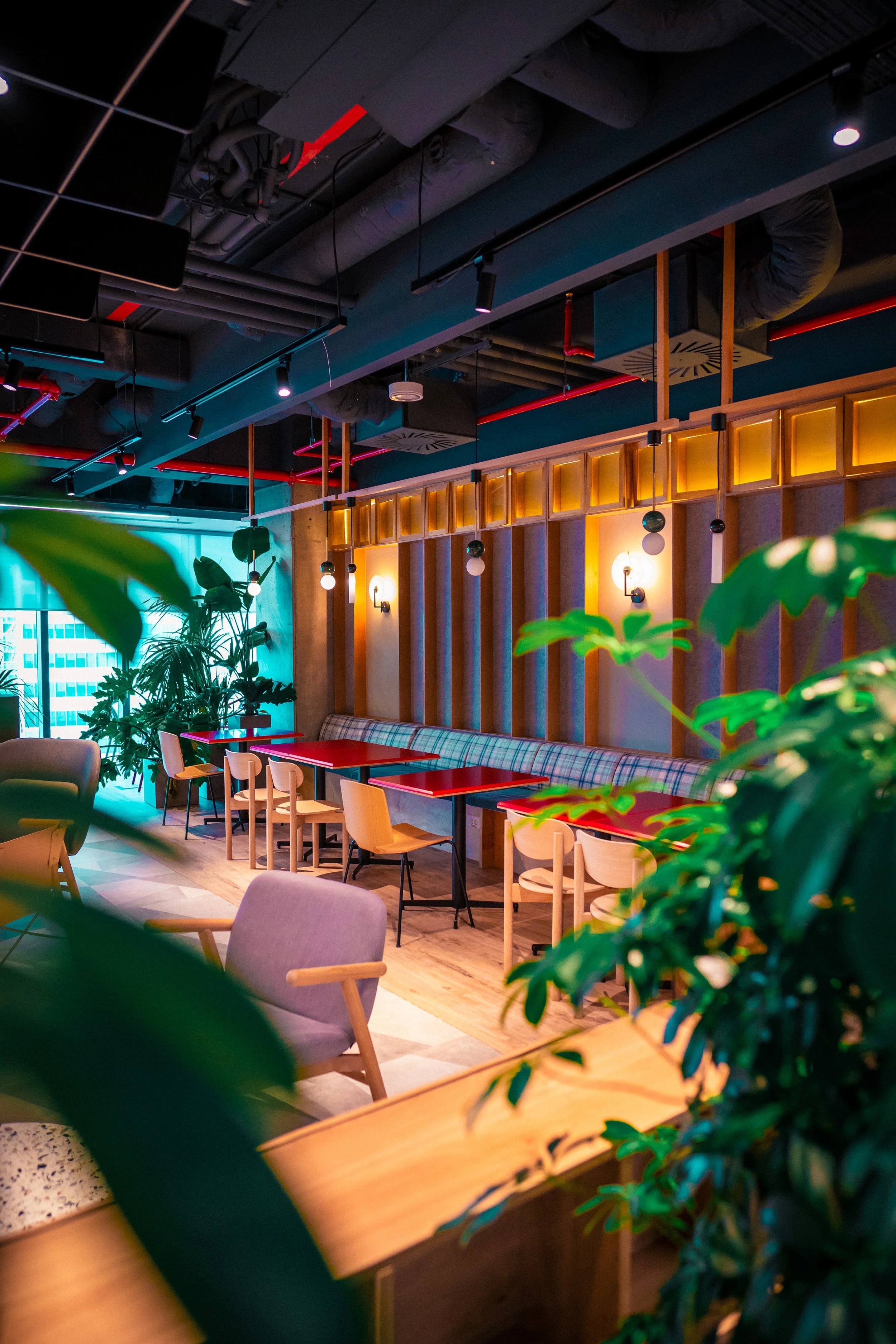 Interior of a modern, stylish cafe or restaurant with colorful lighting, green plants, red tables, and wooden chairs.