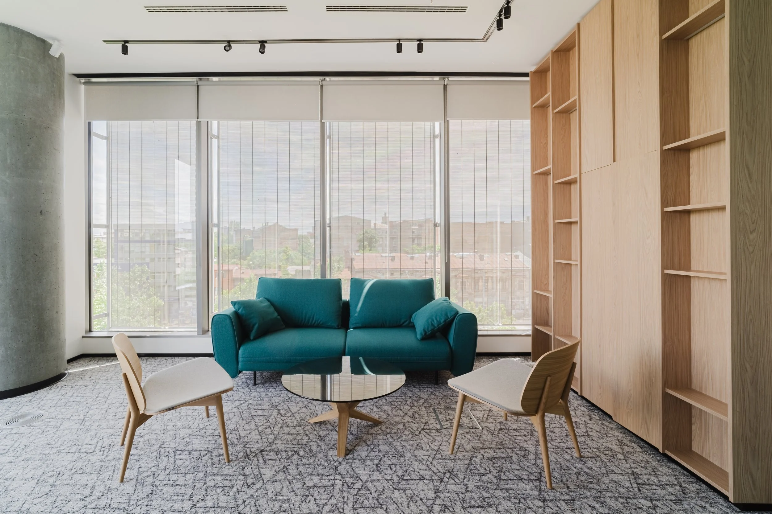 Modern lounge area with a teal sofa, two wooden chairs, a small round glass coffee table, large windows with blinds, and wooden wall cabinets.