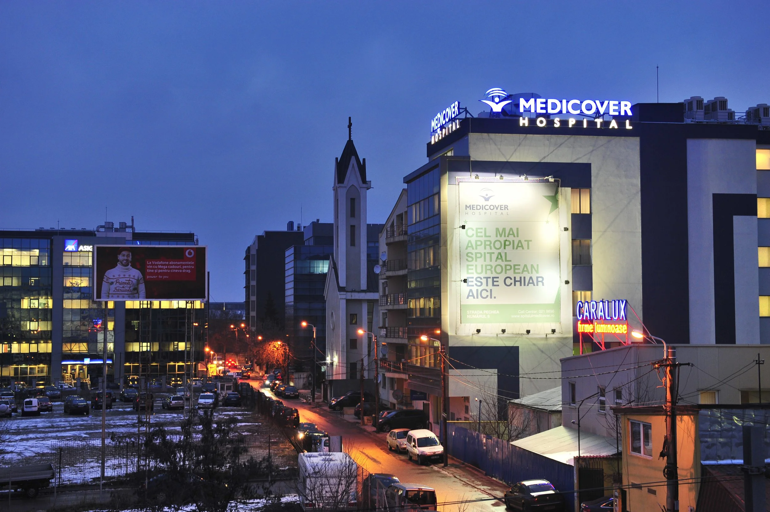 Nighttime cityscape with buildings, cars parked along a street, advertisements on signs, including a cloud-shaped logo for Medicover Hospital and a red-lit sign for Caralux.