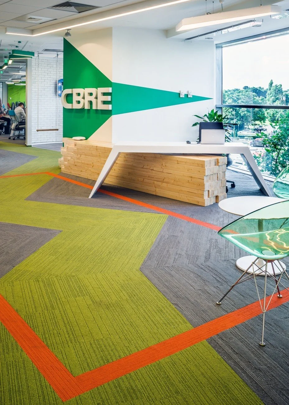 Modern office interior with a reception area displaying the CBRE logo, a white desk with a laptop and plants, colorful carpet tiles in green, gray, and orange, and large windows showing trees outside.