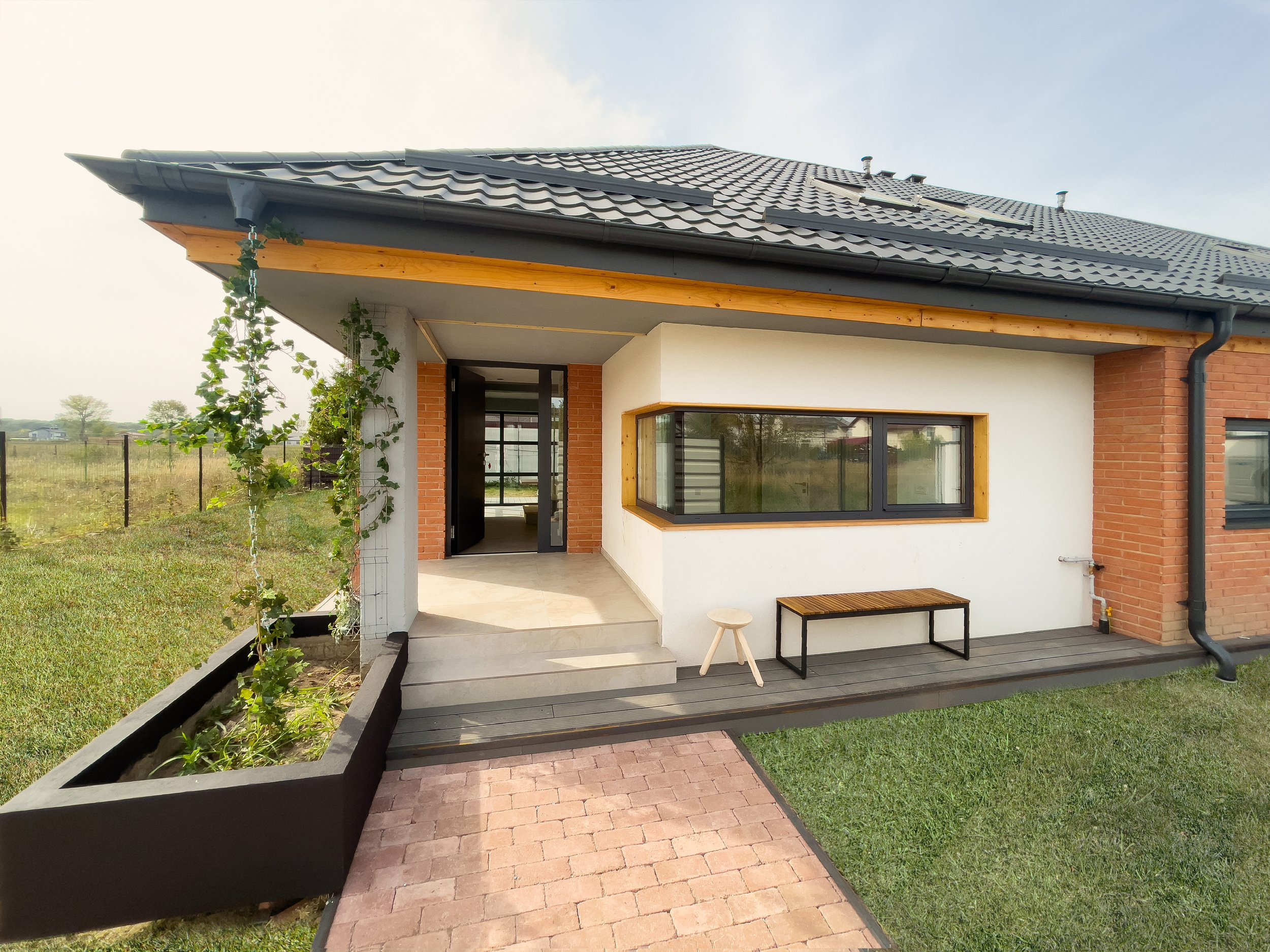 Modern house with brick and white walls, gray roof with solar panels, front porch with stairs, garden, and outdoor seating area.