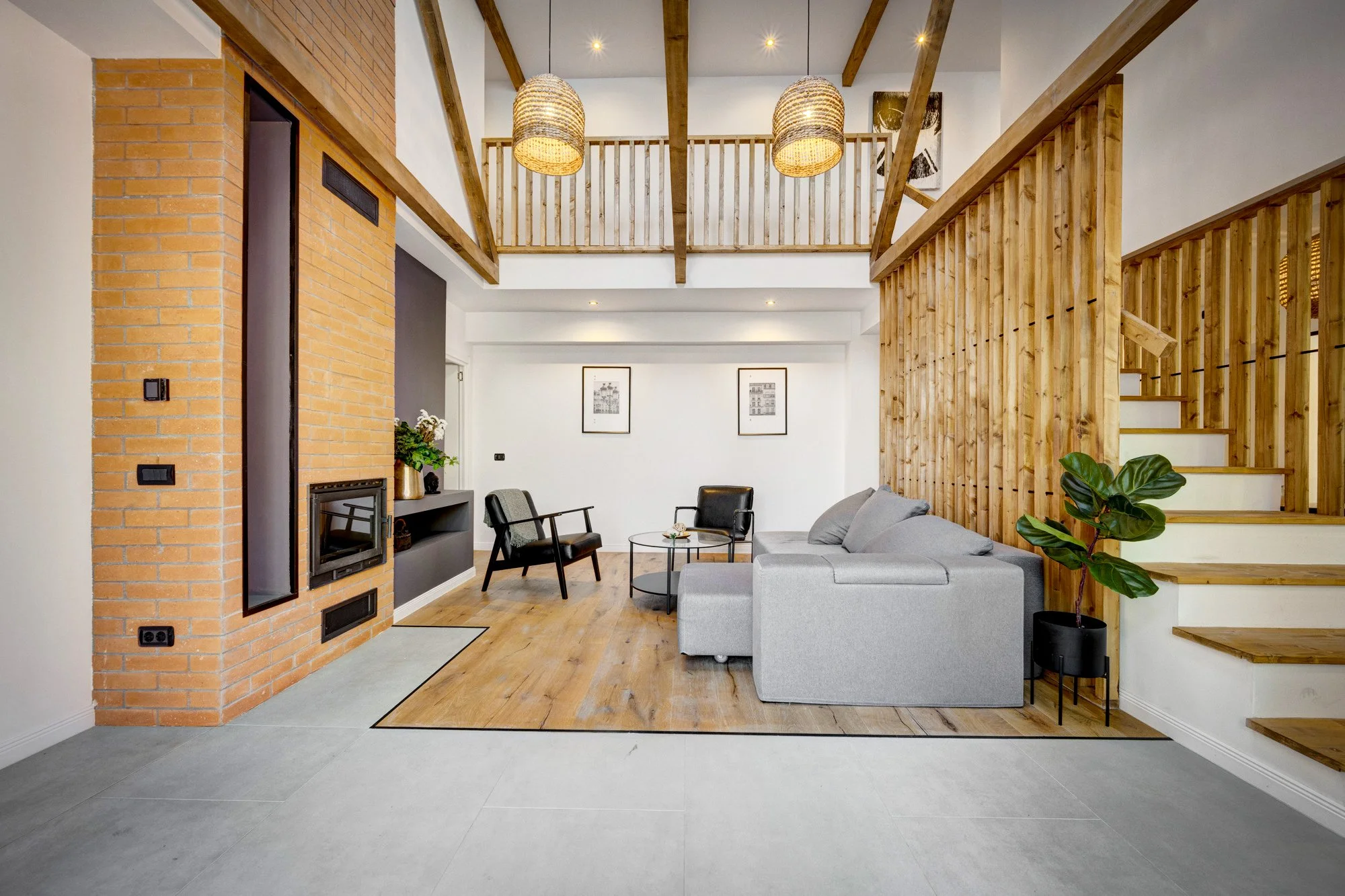 Living room with brick fireplace, gray sofa, black armchair, wooden stairs, and wood accents in ceiling and railing.