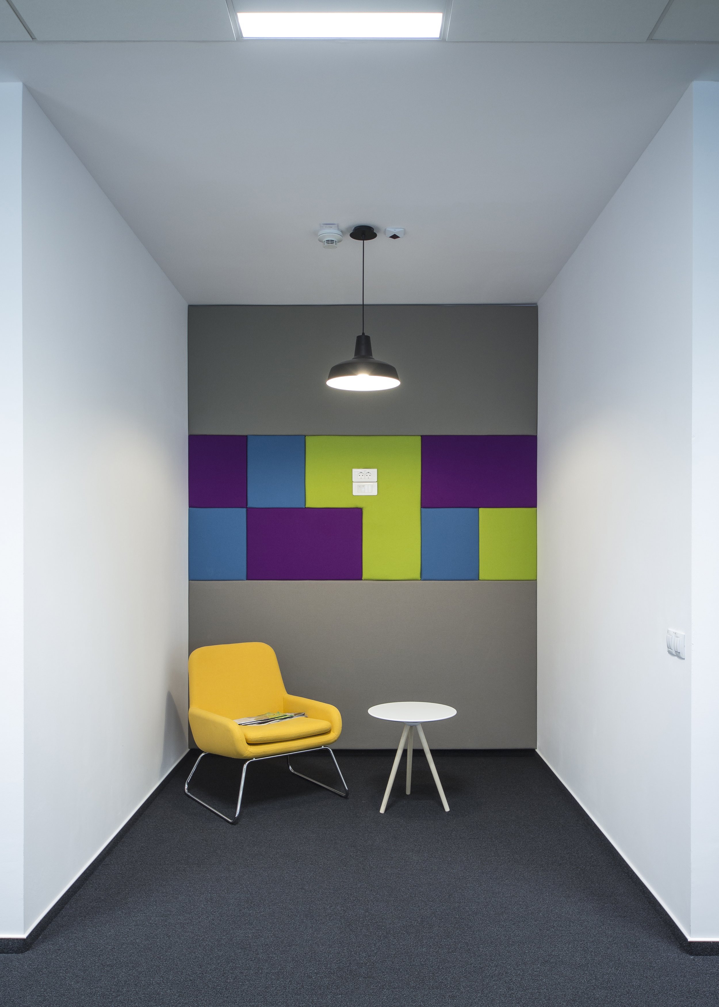 Small, modern waiting area with a yellow armchair, a white round side table, gray carpeted floor, white walls, and a purple, blue, and green abstract wall art piece. A black pendant light hangs from the ceiling.