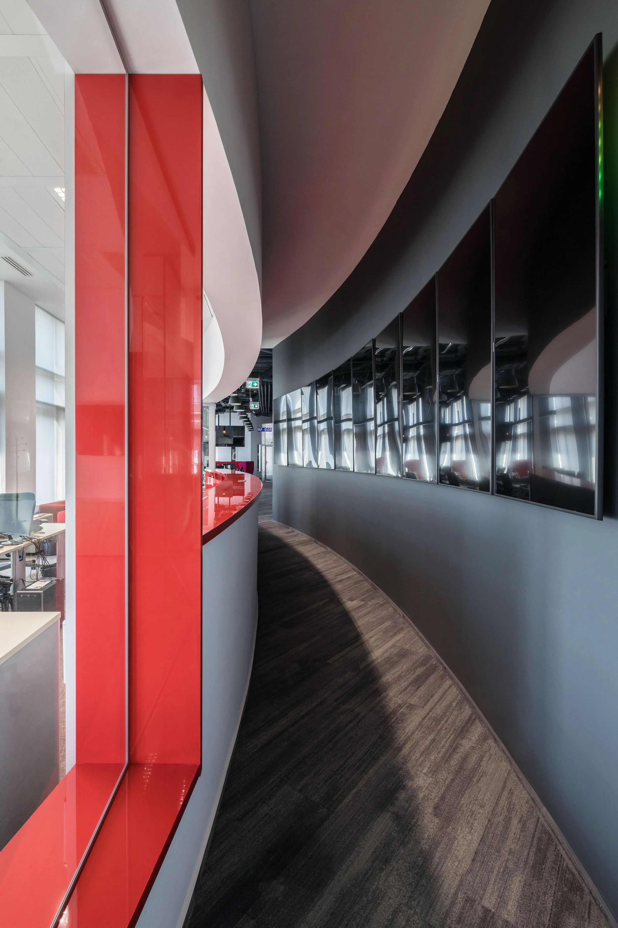 Modern office corridor with curved design, black wall with multiple screens, and a large window with red accents on the left side.