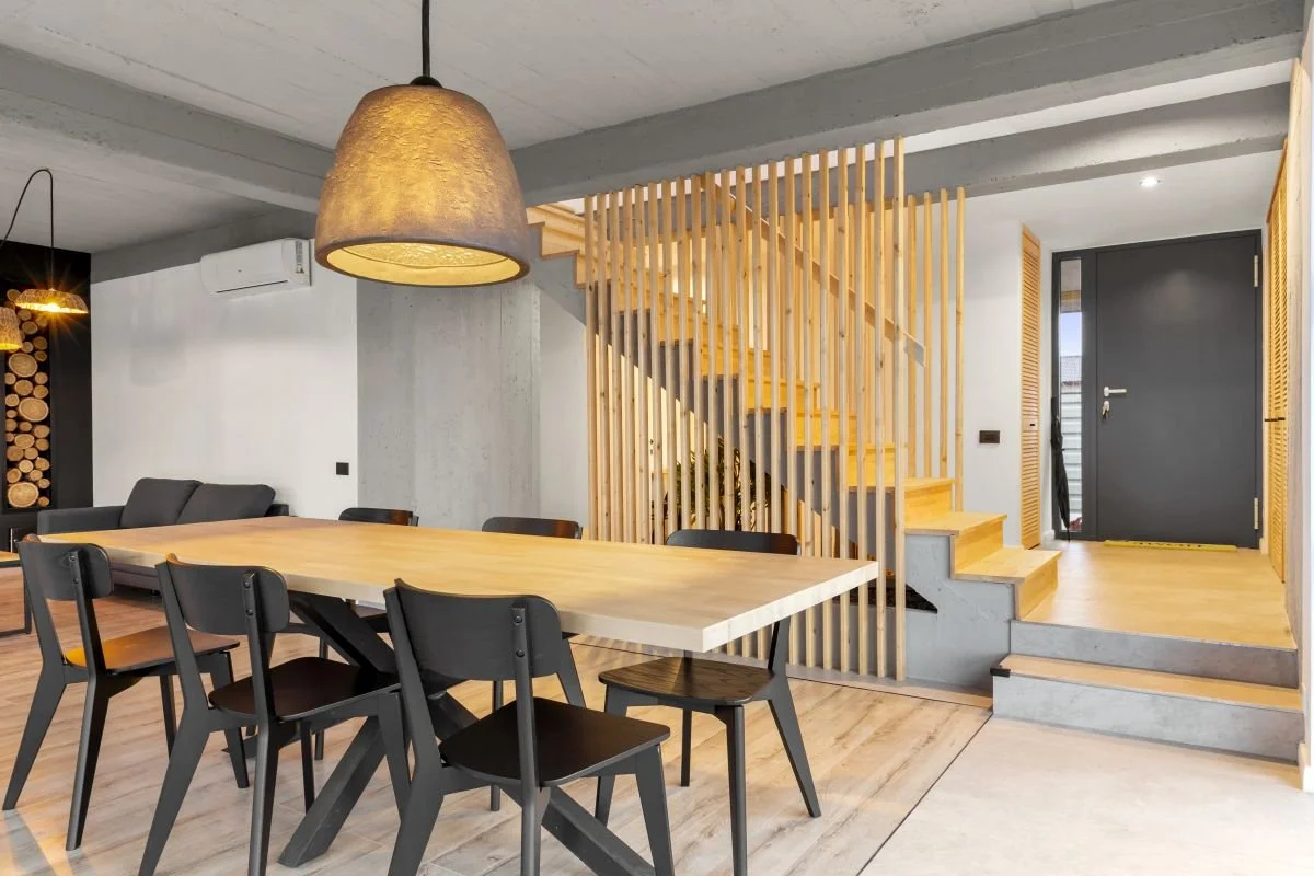 Modern dining room with a large wooden table, black chairs, and a wooden staircase separated by vertical wooden slats, with a front door in the background.