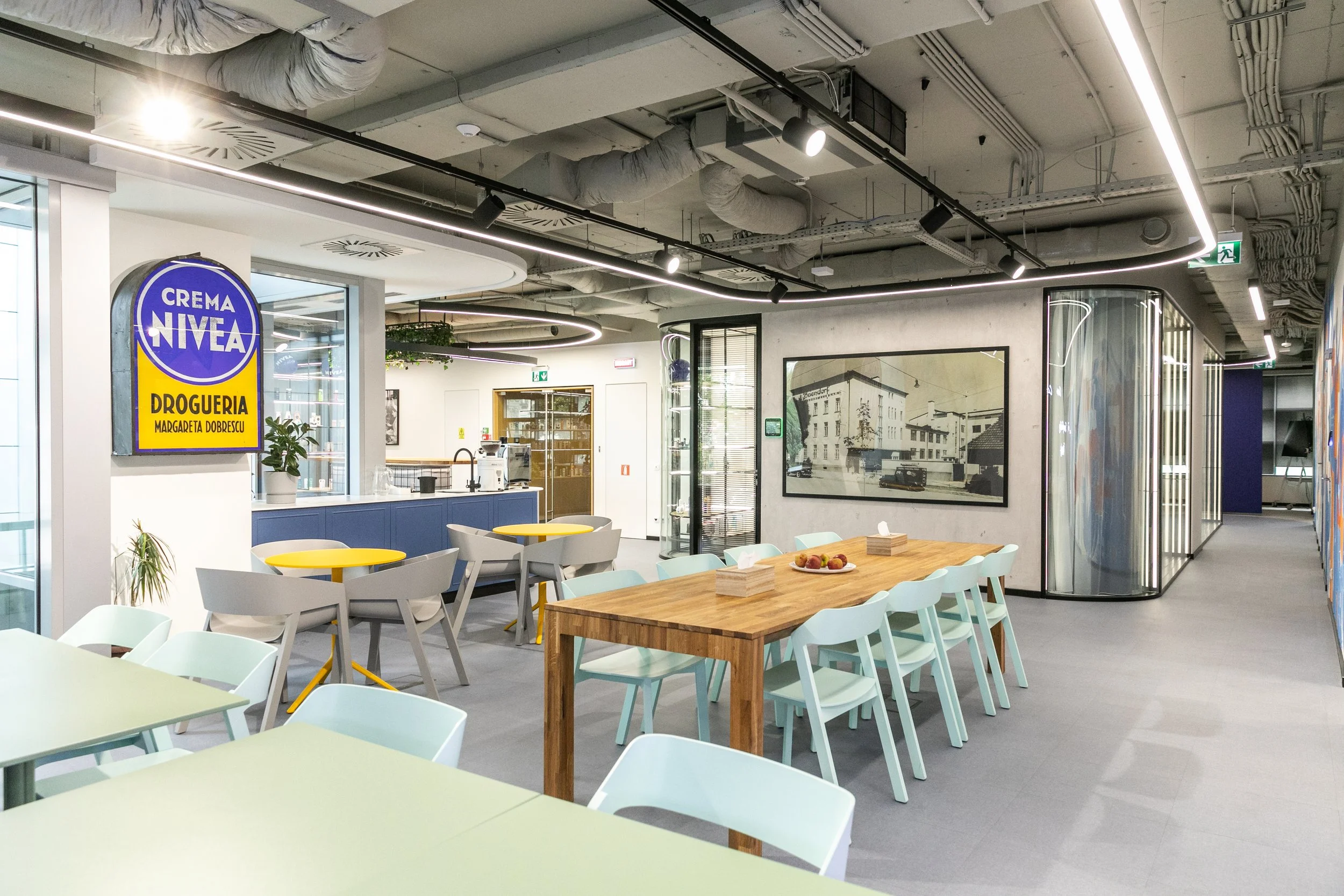 Modern office break room with light-colored tables and chairs, a large framed black-and-white photograph on the wall, a small kitchen area with a blue counter, and a colorful sign that reads 'Crema Nivea Drogueria Magdagueta Dobrescu.'
