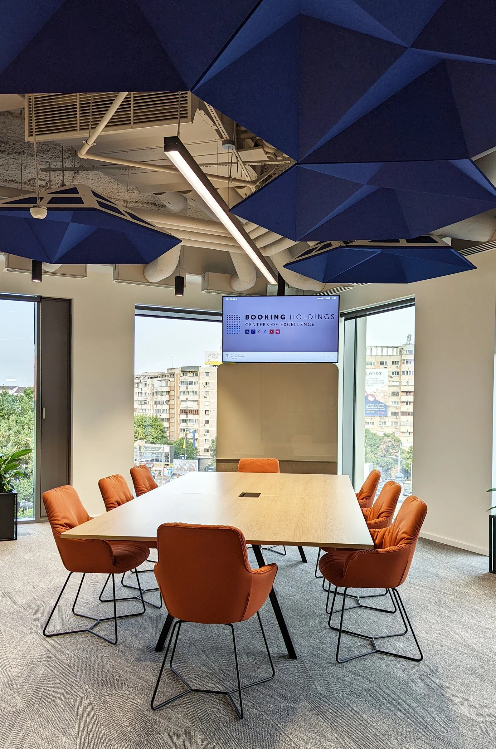 Modern conference room with a wooden table surrounded by orange chairs, large windows, ceiling-mounted blue acoustic panels, and a digital display screen showing 'Booking Holdings Centers of Excellence'.