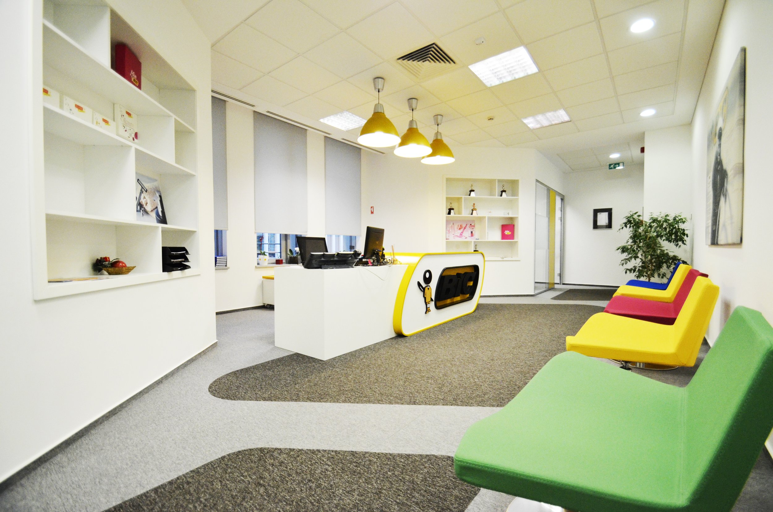 Empty modern office reception area with colorful chairs, white desk, and decorative shelving.