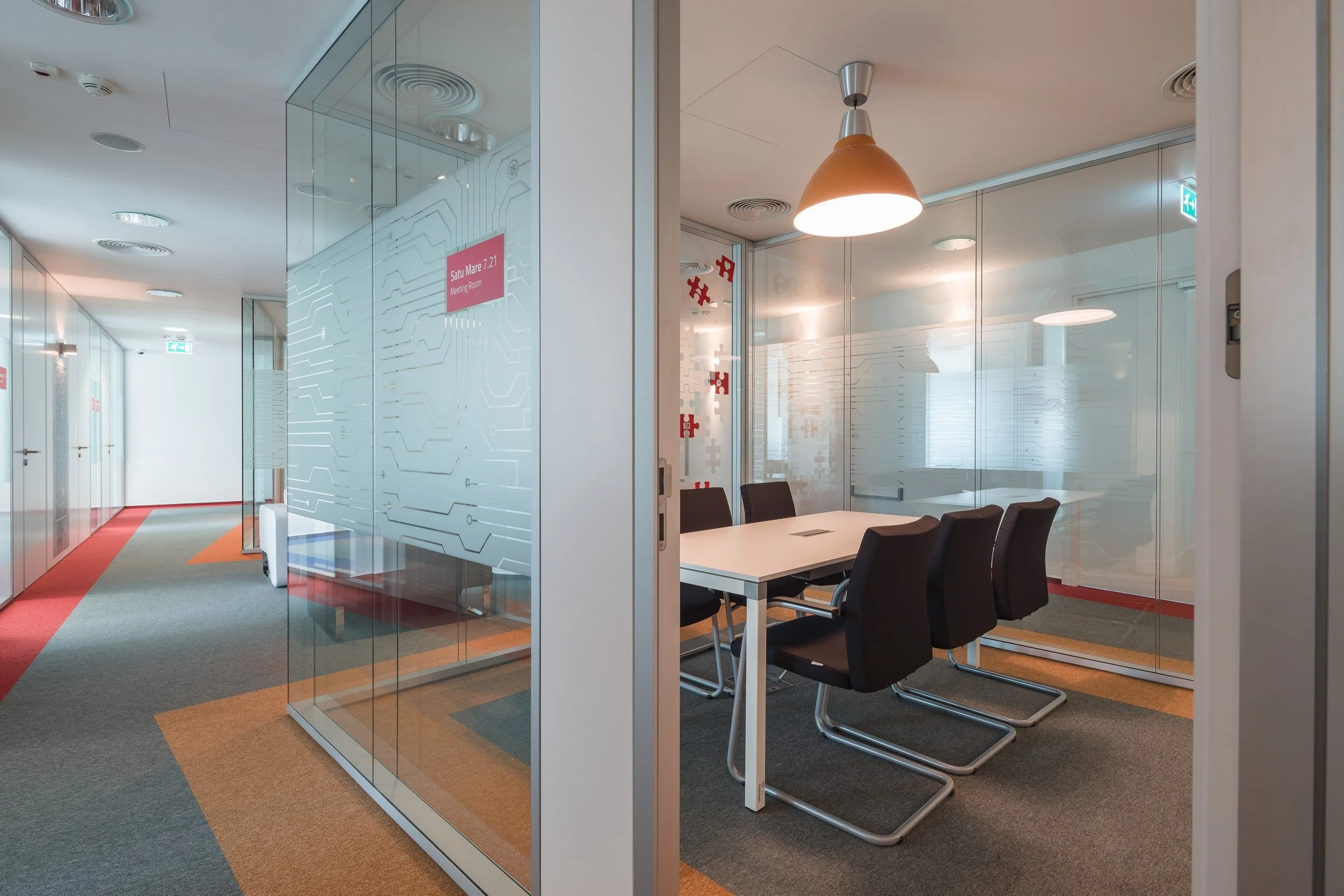 Modern office conference room with a white table, six black chairs, and glass walls. Overhead pendant light and frosted glass with geometric patterns. Adjacent hallway with colorful carpet and white doors.