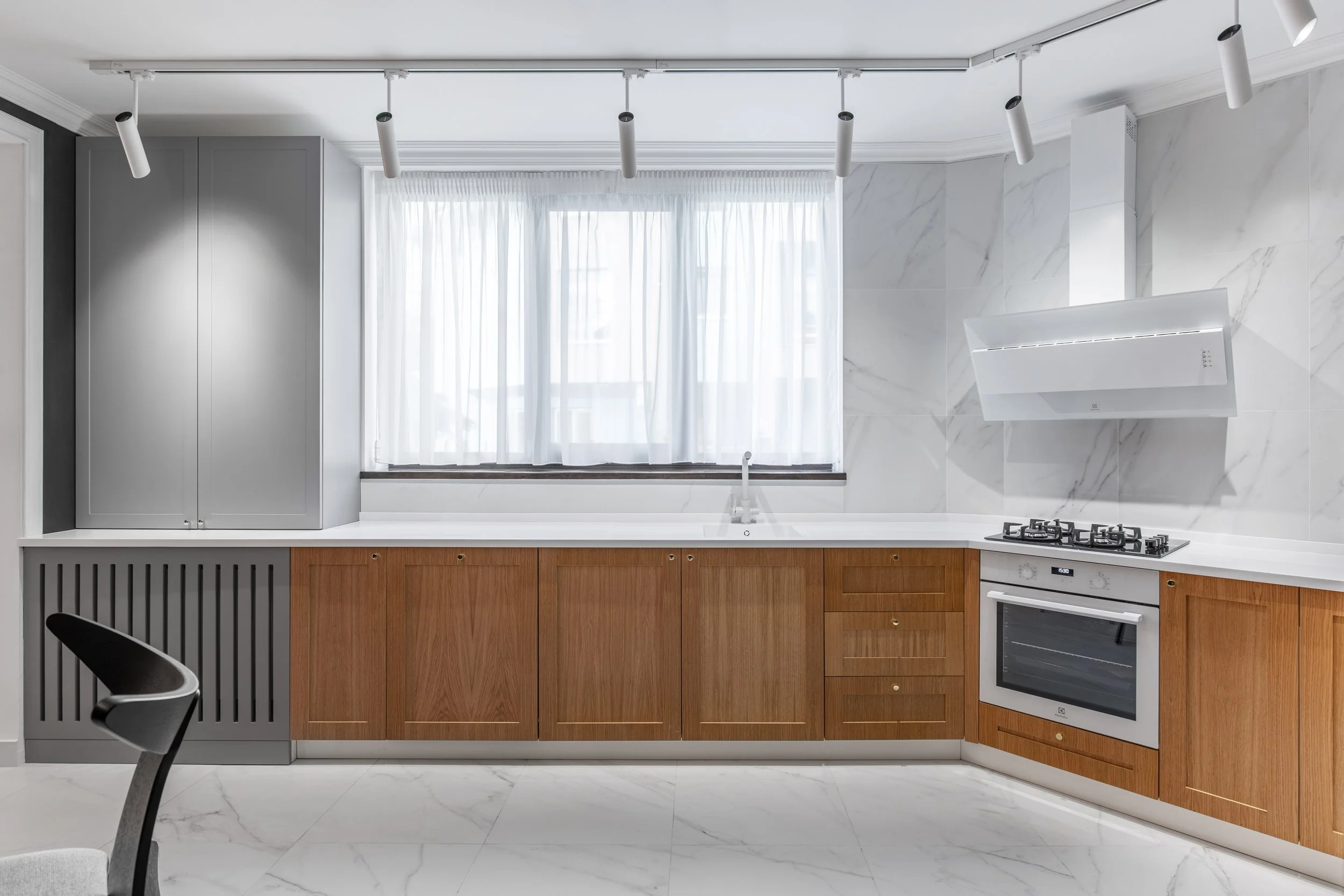 Modern kitchen with wooden cabinets, white countertop, gray upper cabinet, and a stove with an overhead range hood. There's a window with white curtains above the sink, and track lighting on the ceiling.
