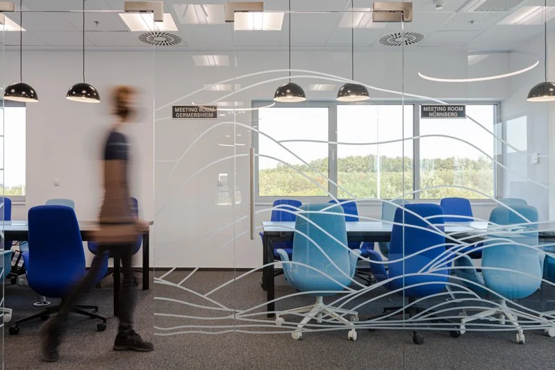 Modern conference room with glass walls, blue office chairs, and large windows showing greenery outside. A person is walking past the room.