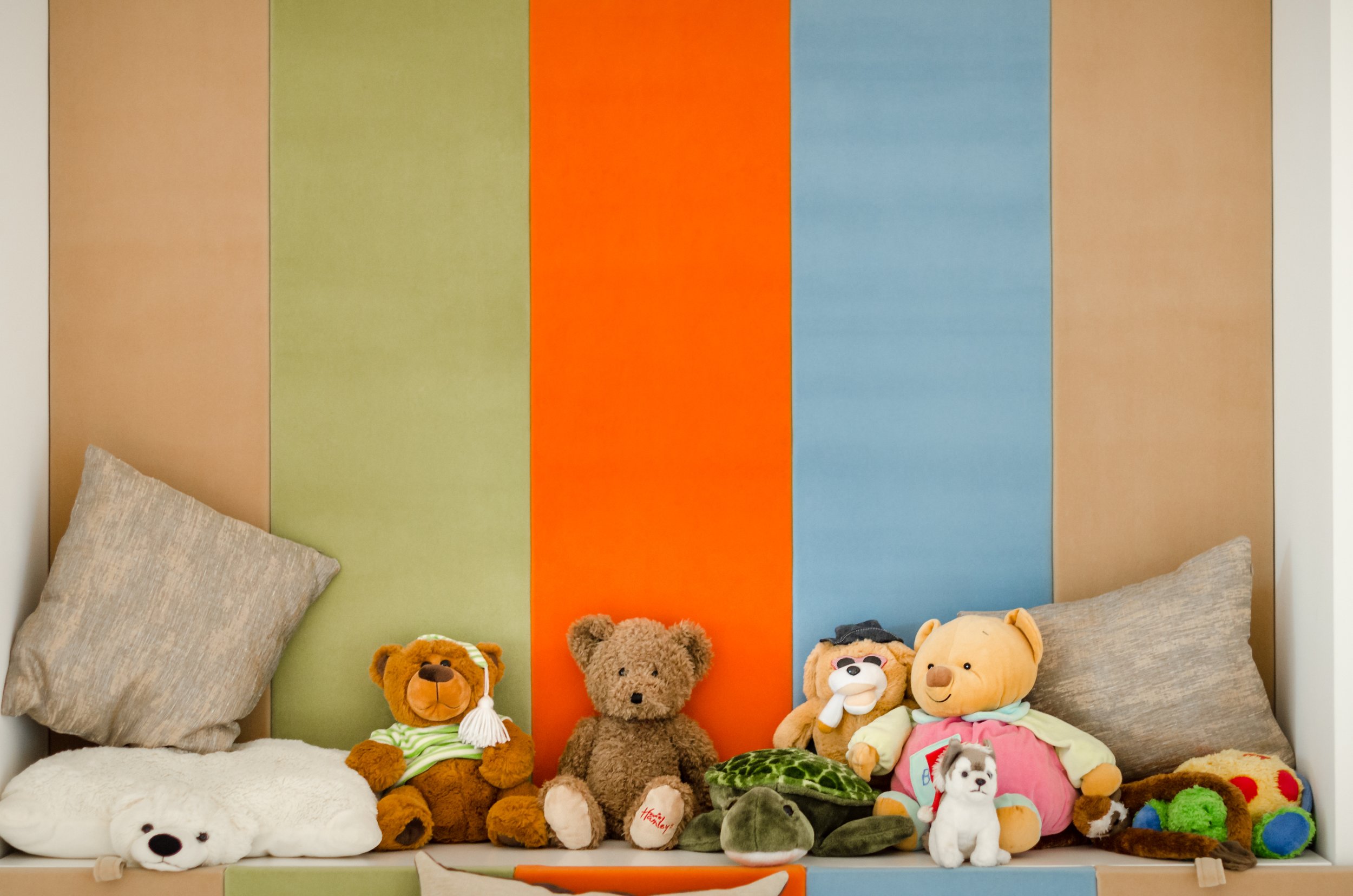 A collection of plush toys and pillows on a bed in front of a colorful striped headboard with vertical sections in green, orange, blue, and beige