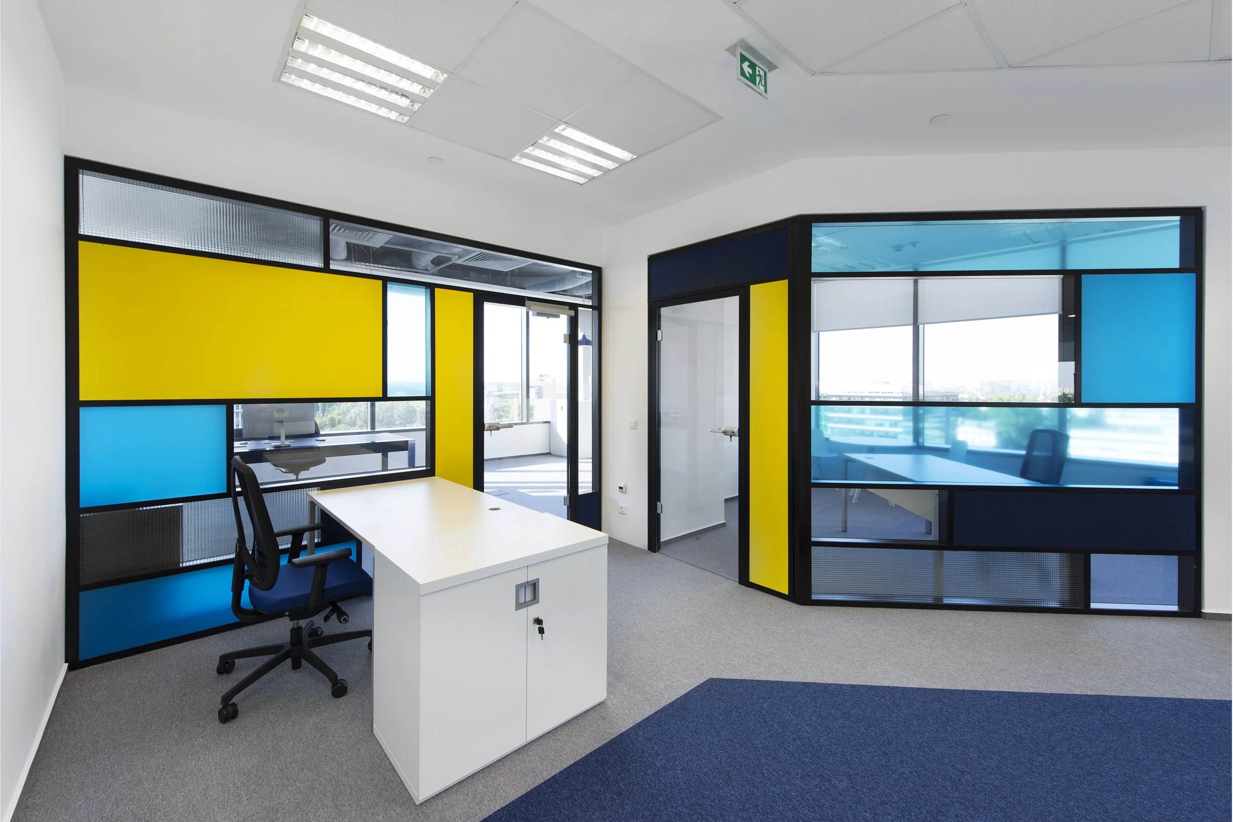 Modern office space with a white desk, black ergonomic chair, colorful glass partition walls in yellow and blue, and large windows allowing natural light, with a carpeted floor.