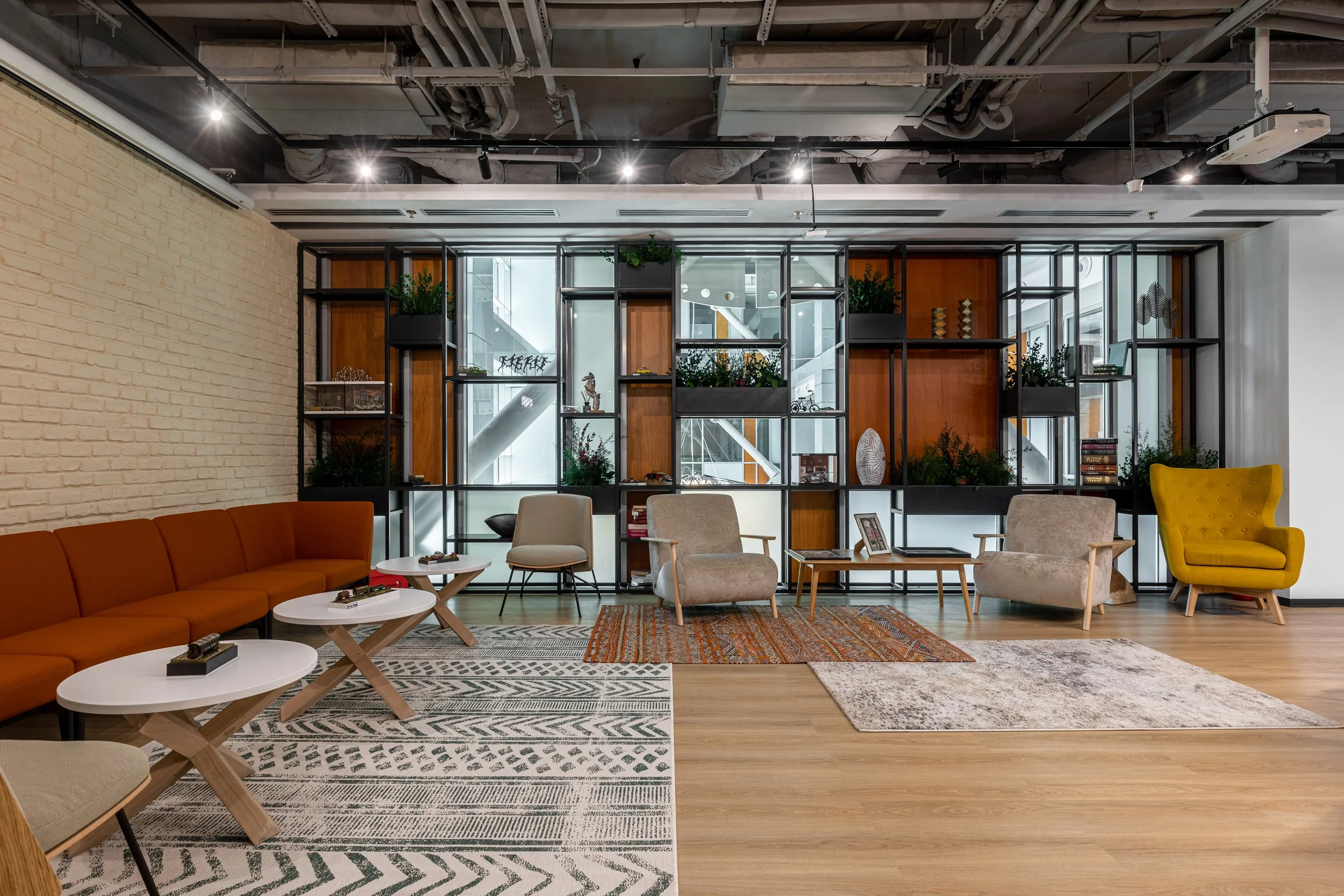 Modern lounge area with a variety of chairs and sofas, a decorative shelving unit behind, hardwood floors, and ceiling with exposed pipes and ducts.