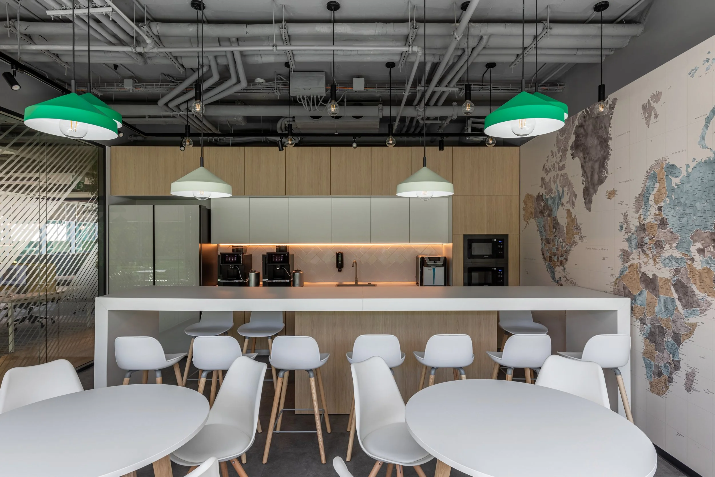 Modern kitchen and dining area with white tables and chairs, bar seating, wood and gray cabinetry, coffee machines, a world map on the wall, pendant lights, and an open ceiling with exposed pipes.