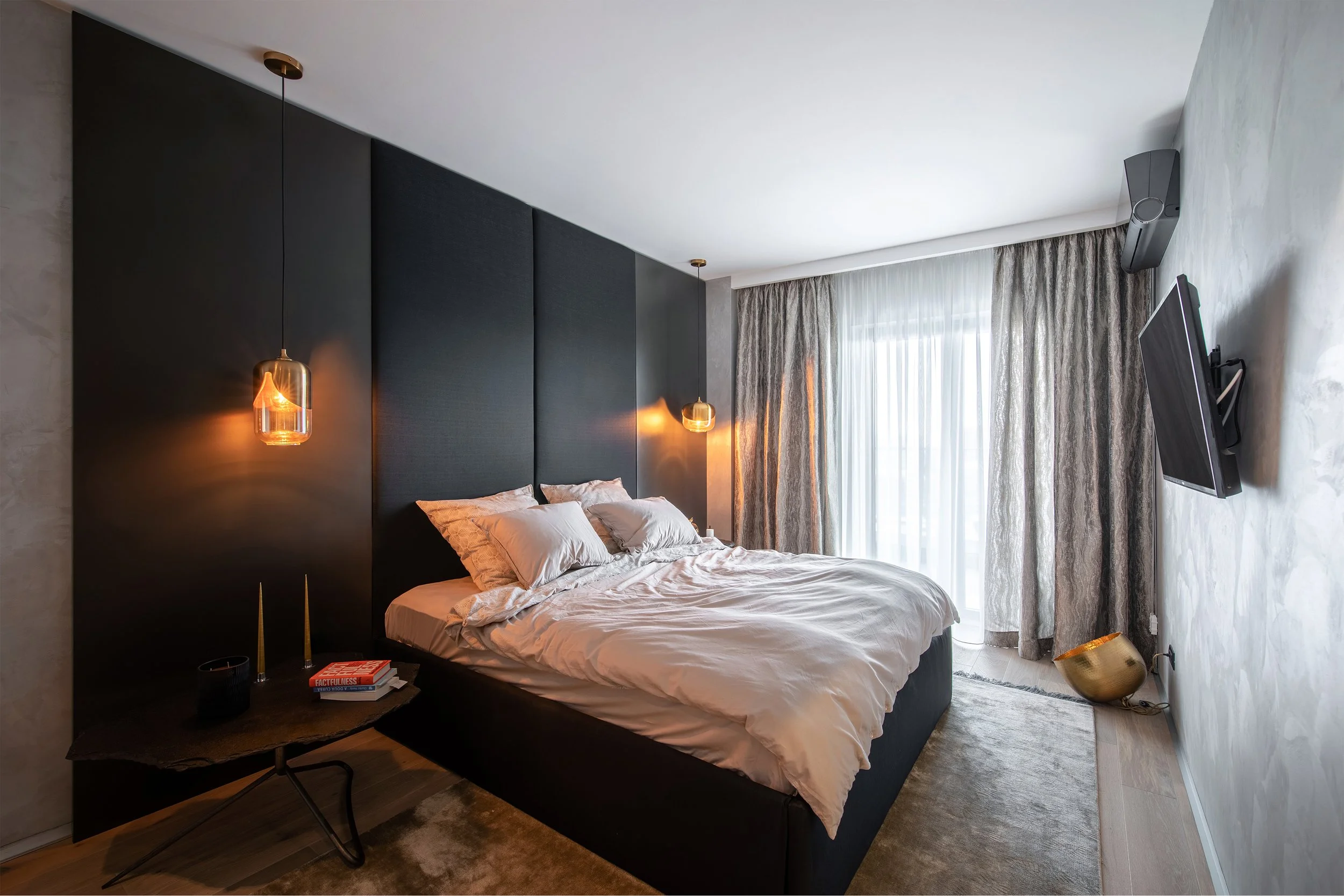 Modern bedroom with a black upholstered headboard, white bedding, and bedside lamps. A side table with books and candles, a textured rug, a wall-mounted TV, and large curtains covering a window or balcony door.