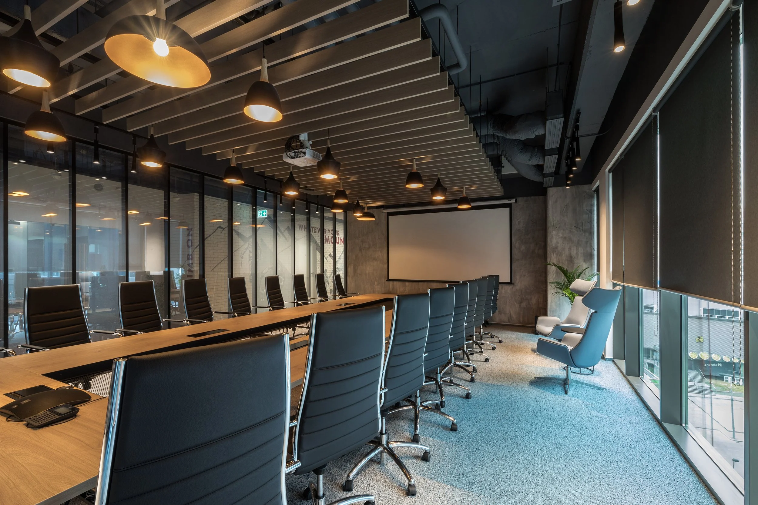 Modern conference room with a long wooden table, black leather chairs, a white executive chair, and a large projection screen. The room has glass walls, large windows with black curtains, and ceiling-mounted lighting fixtures.