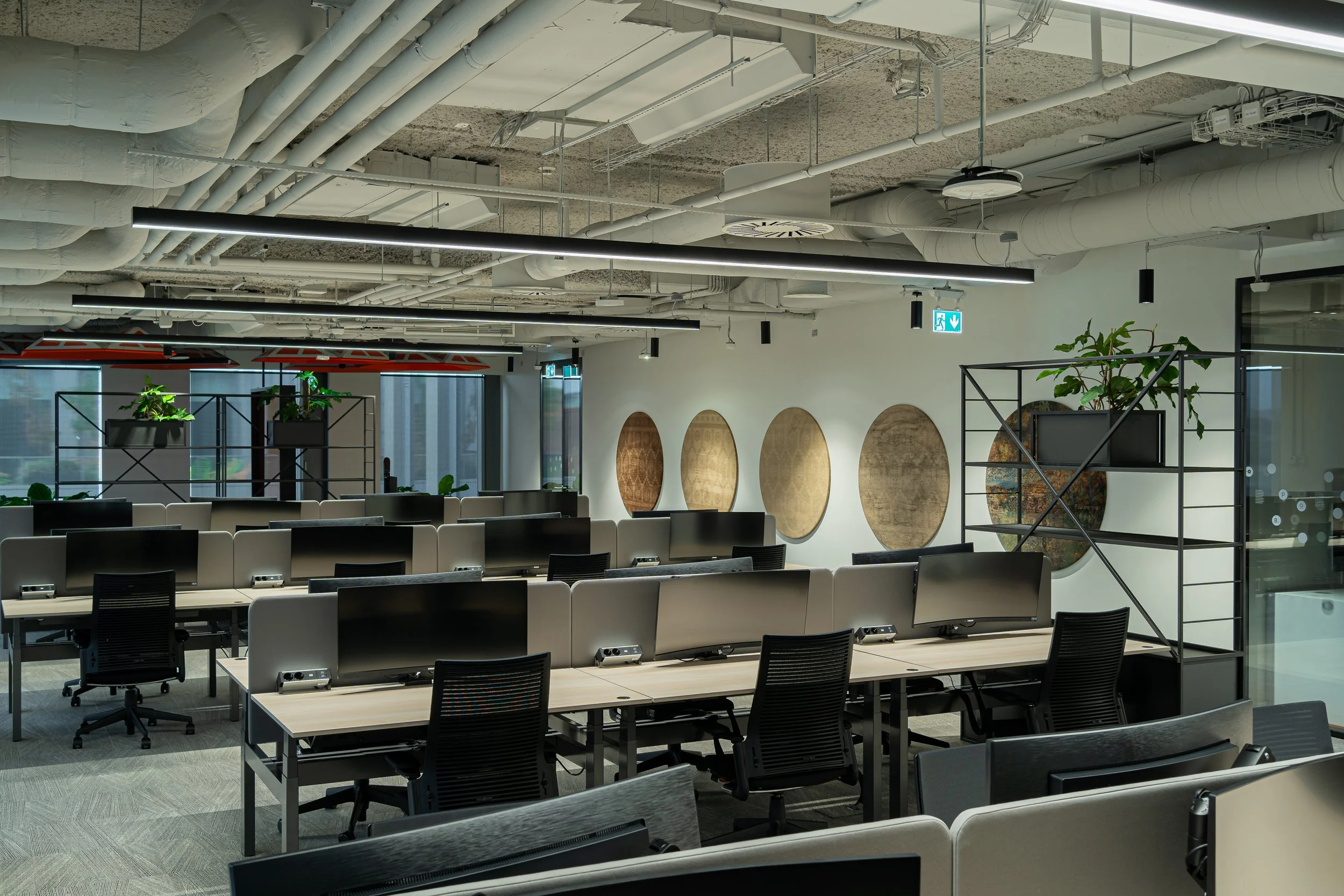 Modern office space with rows of workstations, black office chairs, and divided computer desks. The ceiling features exposed pipes and ducts, and there are round wall decorations and green plants on shelving units.