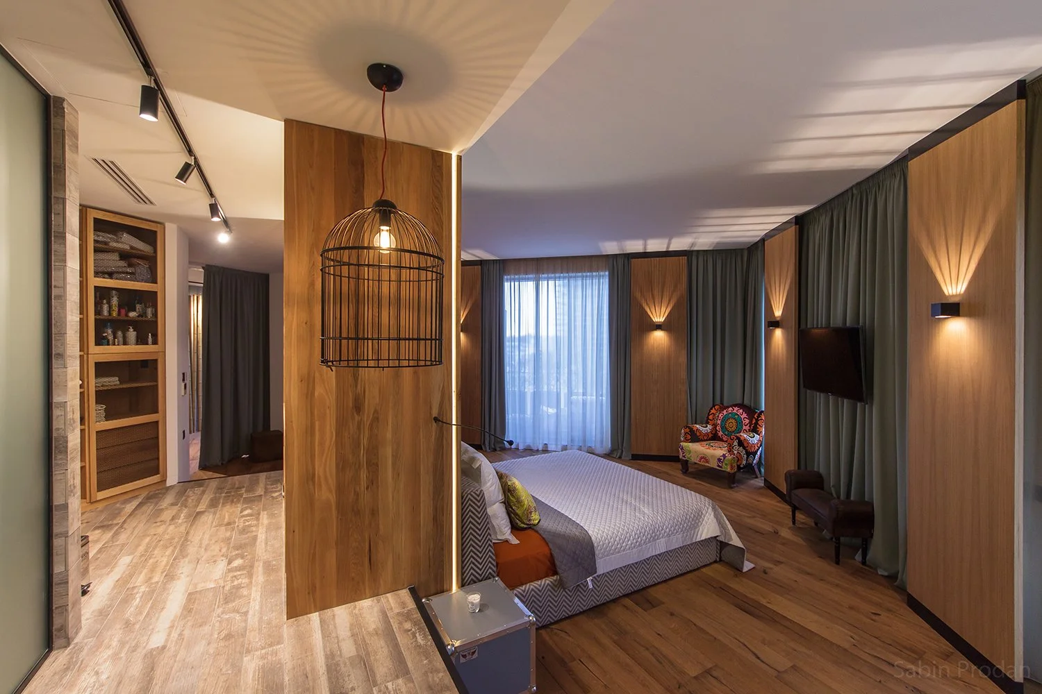 Modern bedroom with wooden wall panels, a large window with sheer curtains, a patterned armchair, a wall-mounted flat-screen TV, and wall sconces emitting warm light.