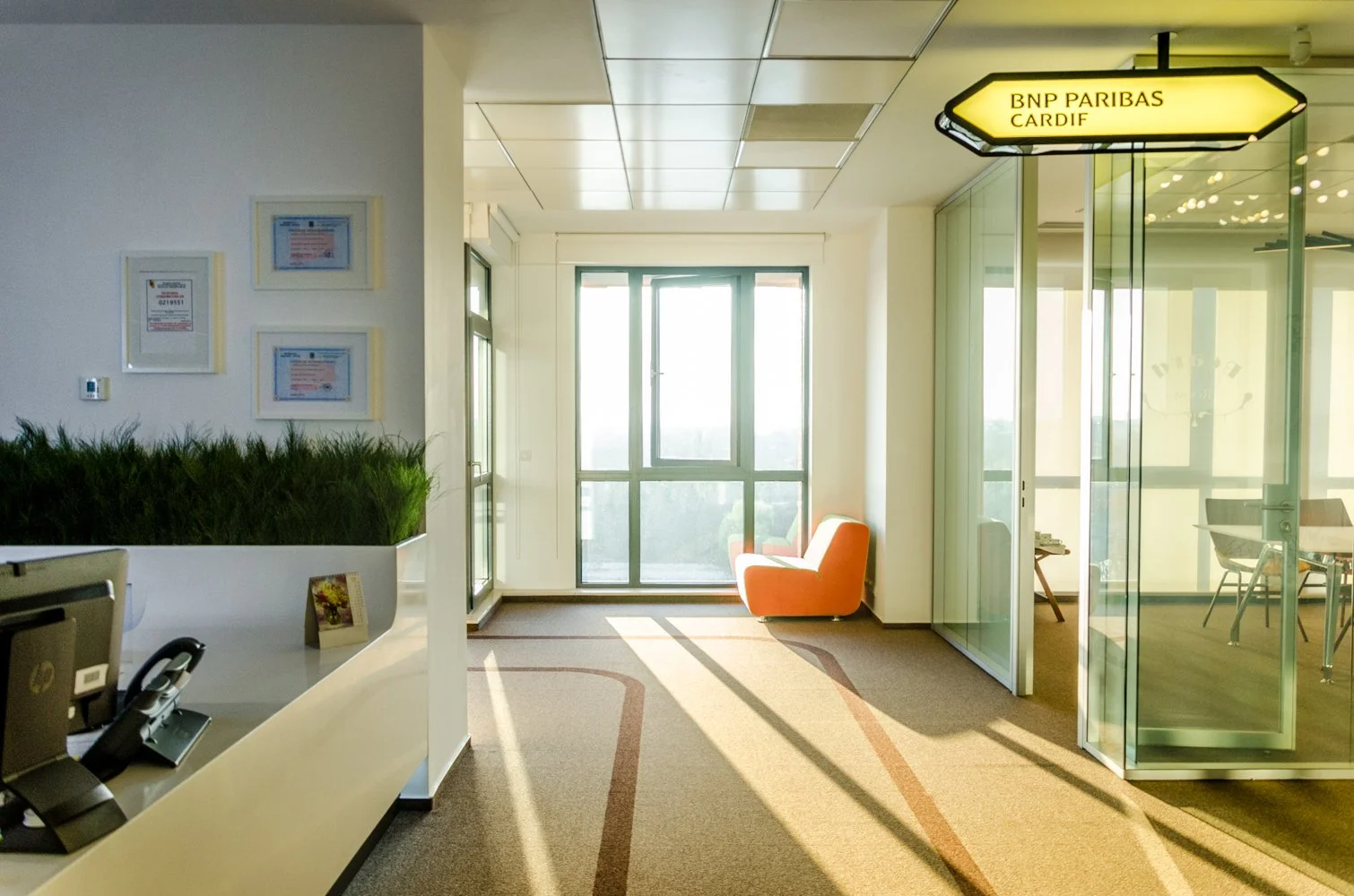 Bright office lounge with orange armchair near large window, glass enclosed meeting room to the right, and a reception desk with a telephone and plants on the left.
