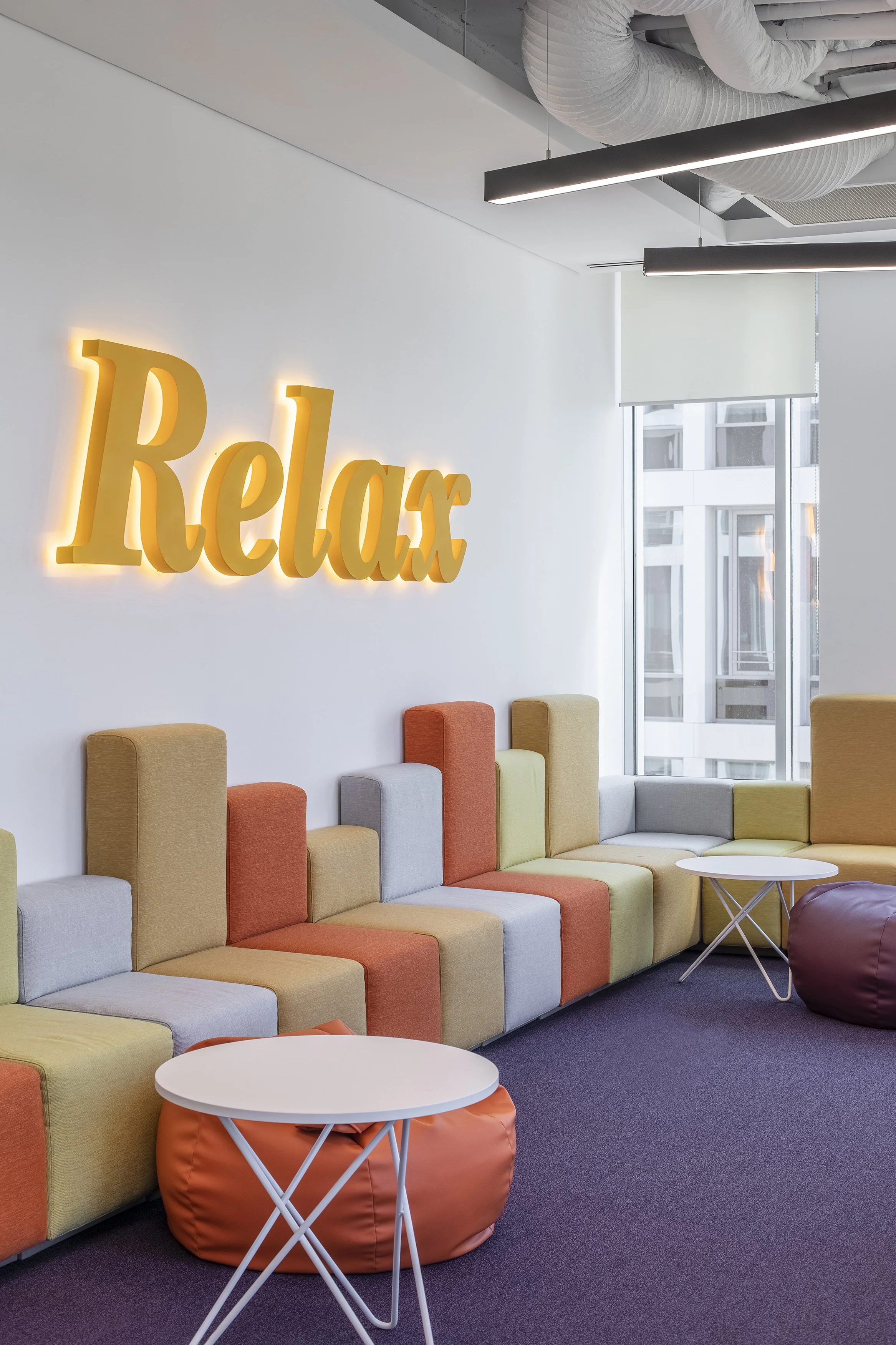 Modern lounge area with multicolored cushioned seats, small white tables, and a large illuminated 'Relax' sign on the wall, near a window with a city view.