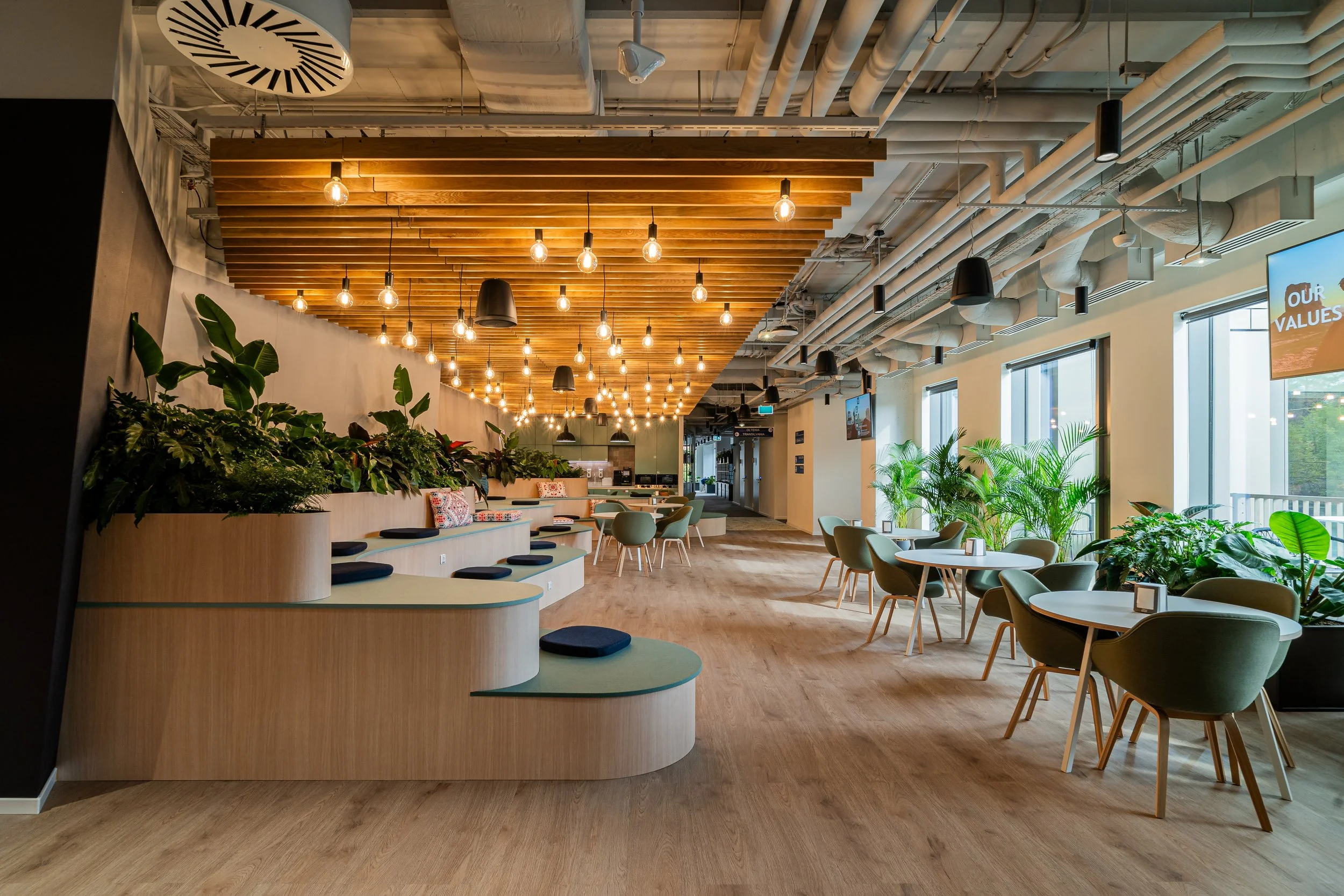 Modern office interior design with green chairs and round tables near large windows, wooden flooring, a ceiling with exposed pipes, and hanging light bulbs, decorated with lots of green plants.