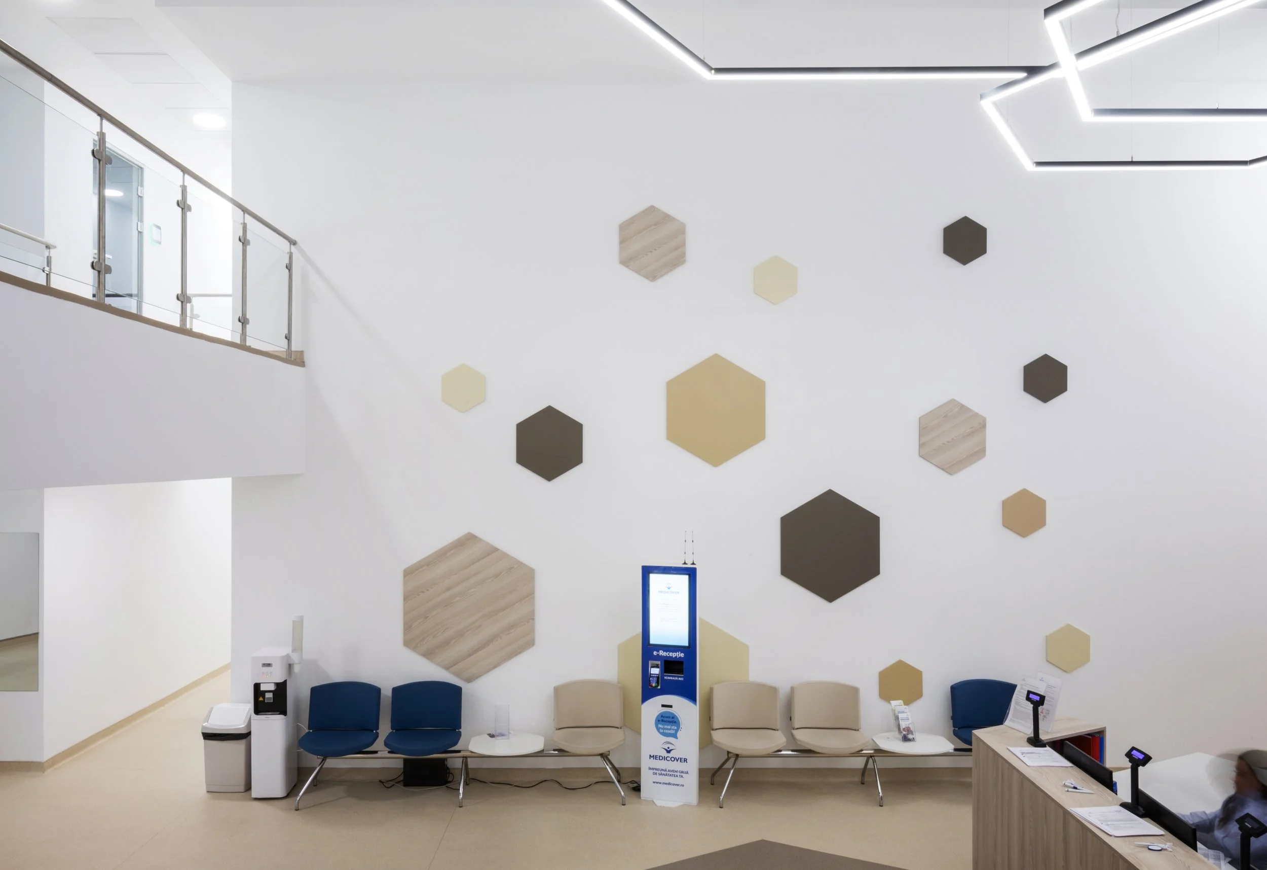 Waiting area of a medical facility with beige and blue chairs, a digital check-in kiosk, a reception desk on the right, and wall decorations of hexagonal panels in neutral colors.