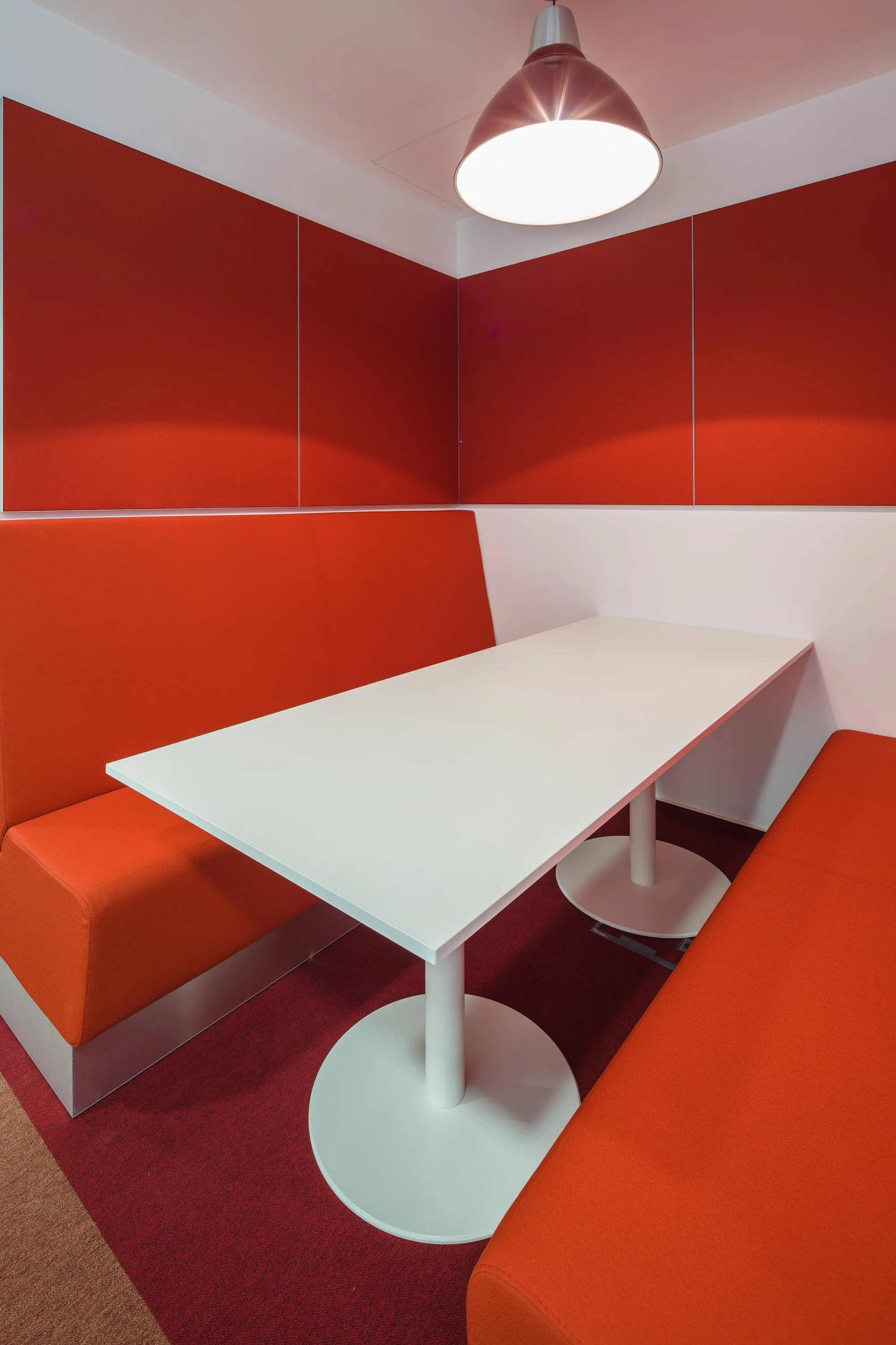A small, modern dining nook with a white table, orange cushioned bench seats, and red wall panels, lit by a metallic pendant light.