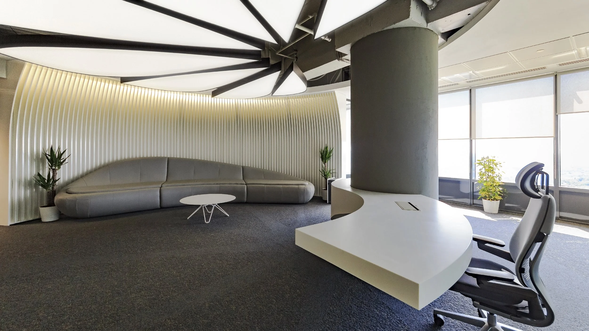 Modern office space with a curved gray sofa, white wall with vertical ridges, small white round coffee table, and potted plants. A large white desk with an ergonomic chair and windows with blinds letting in sunlight.