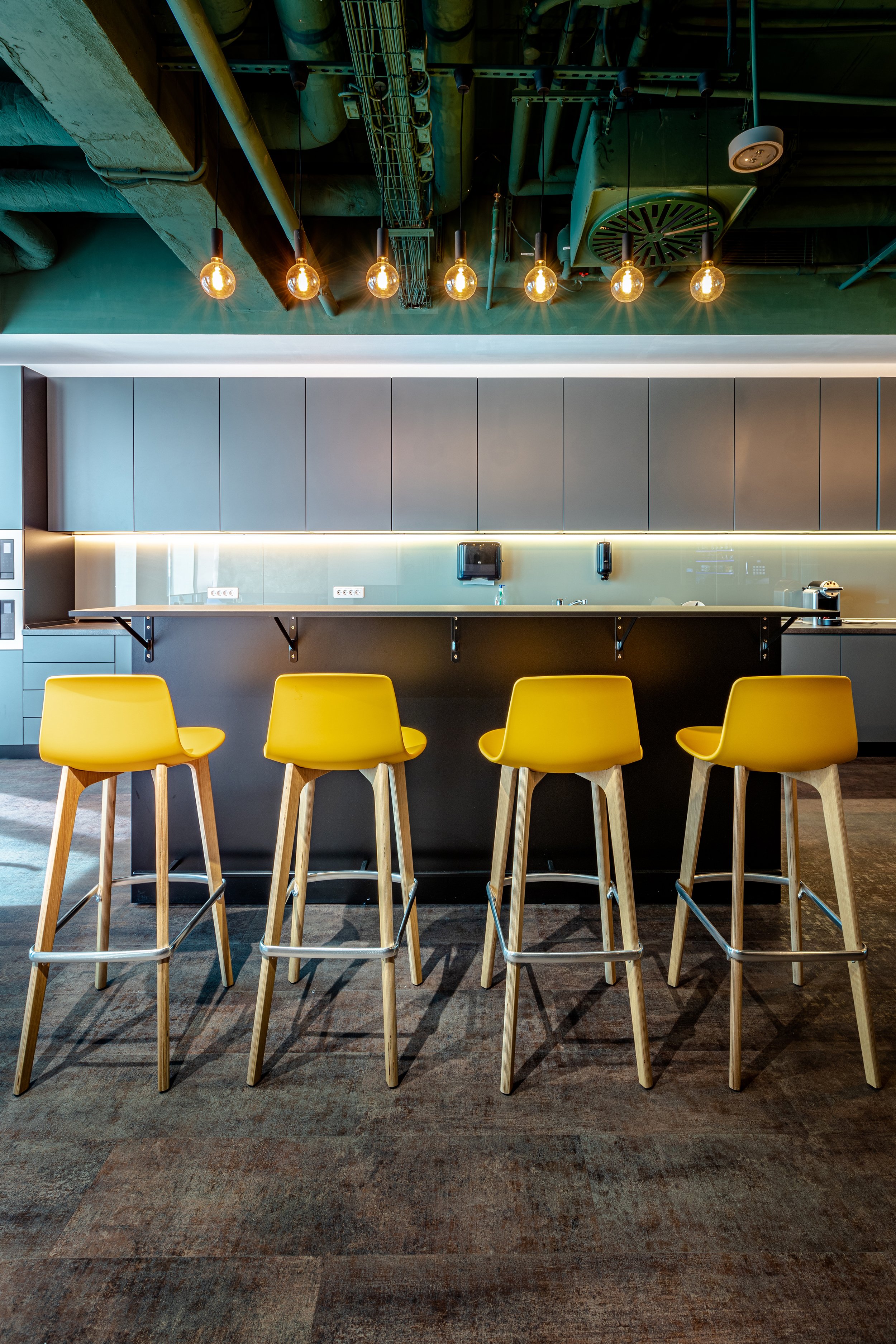 Modern kitchen with a black counter, gray upper cabinets, yellow bar stools, and industrial ceiling with exposed pipes and hanging Edison bulbs.