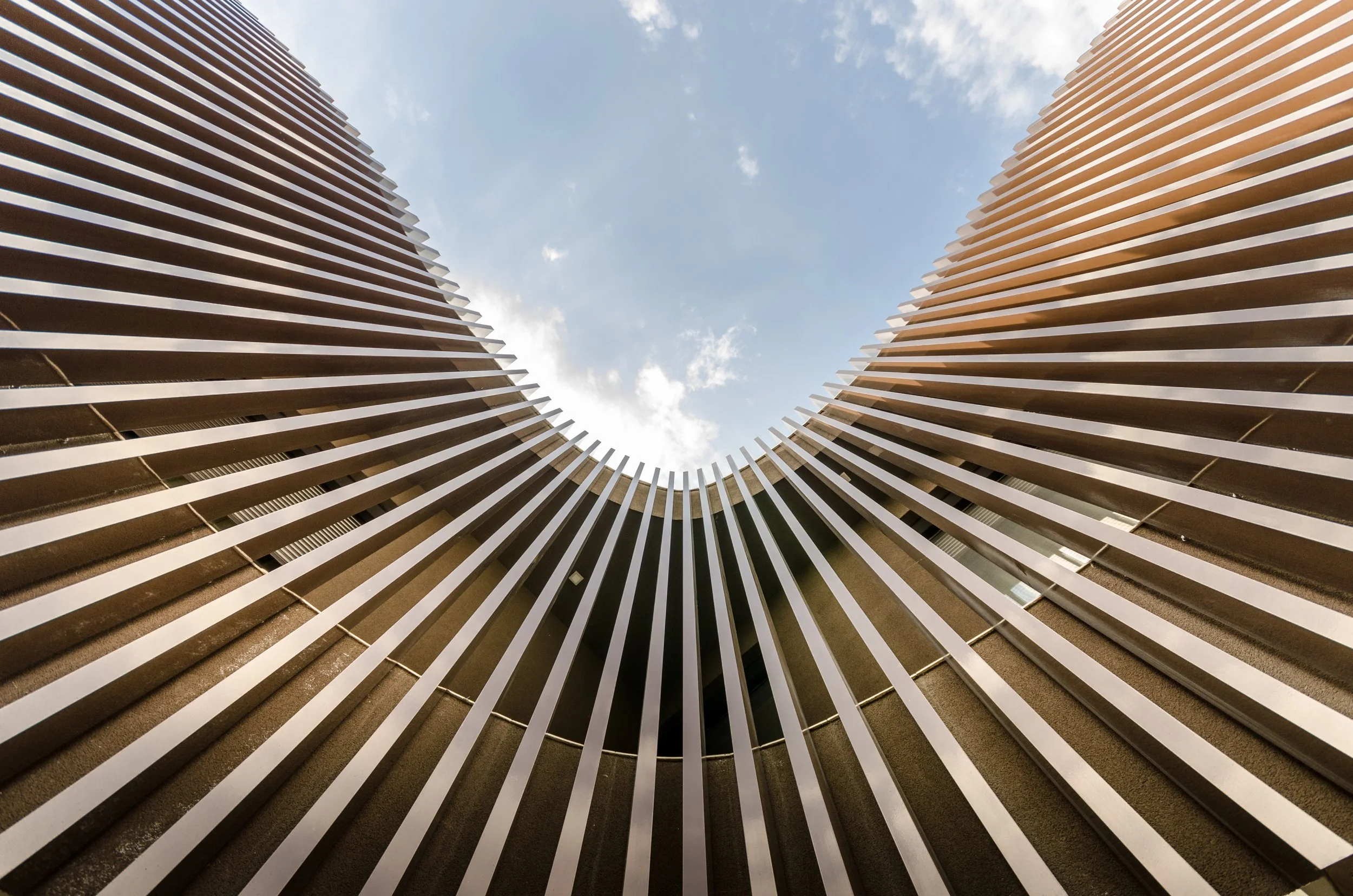 Looking up at a modern building with vertical metal slats forming a rounded opening, with a blue sky and clouds visible.