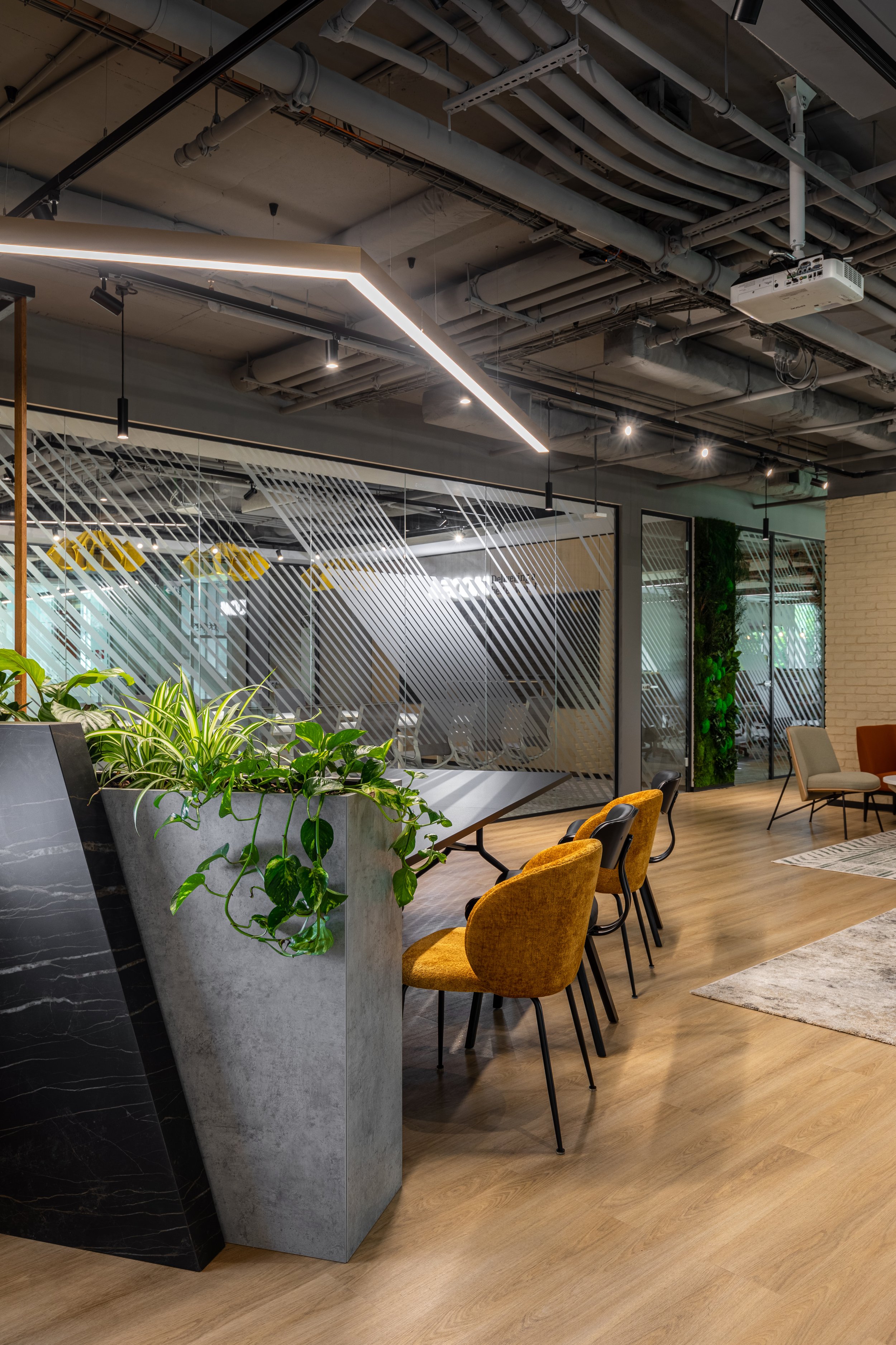 Modern office lounge area with yellow chairs, a concrete planter with green plants, glass walls with diagonal striped designs, exposed ceiling pipes, and wooden flooring.