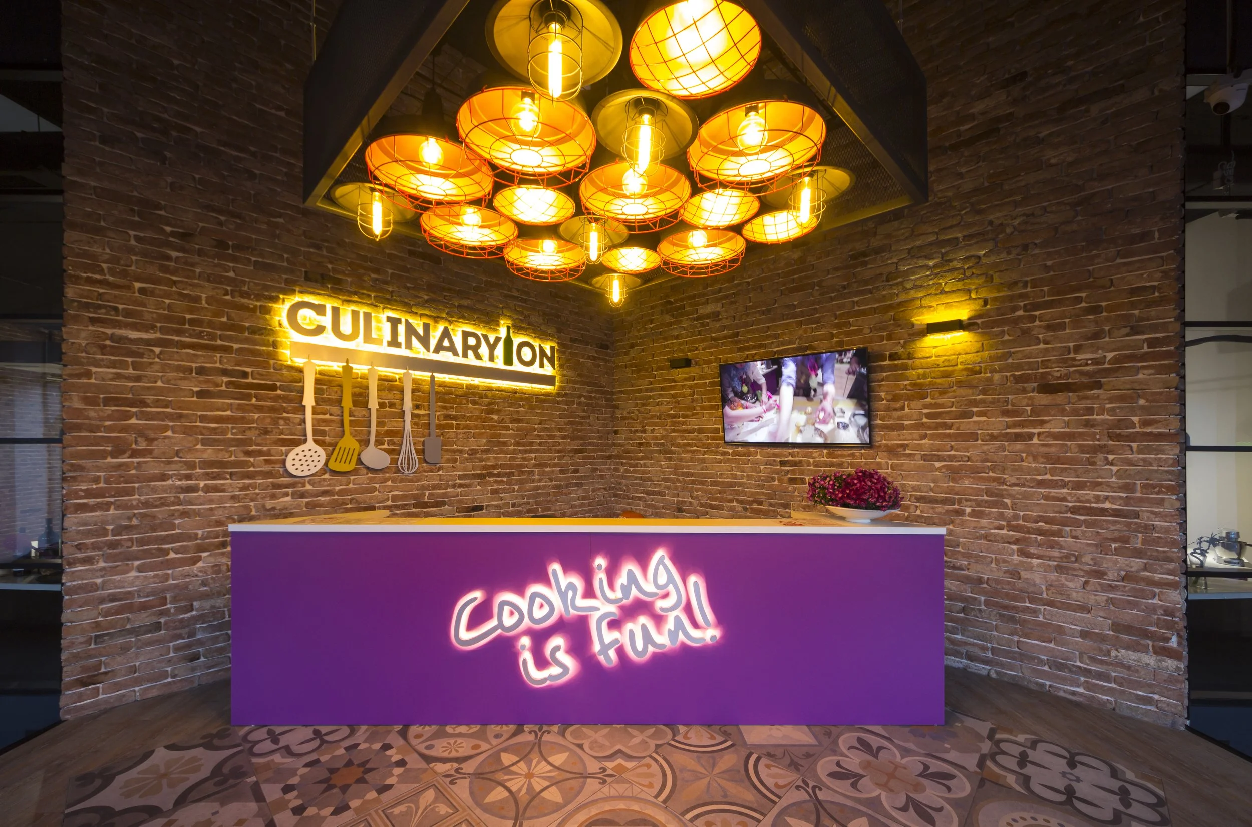 A colorful cooking studio with a brick wall, a neon sign that says 'cooking is fun!', hanging orange and yellow lamp lights, a purple counter, and kitchen utensils decoration on the wall.
