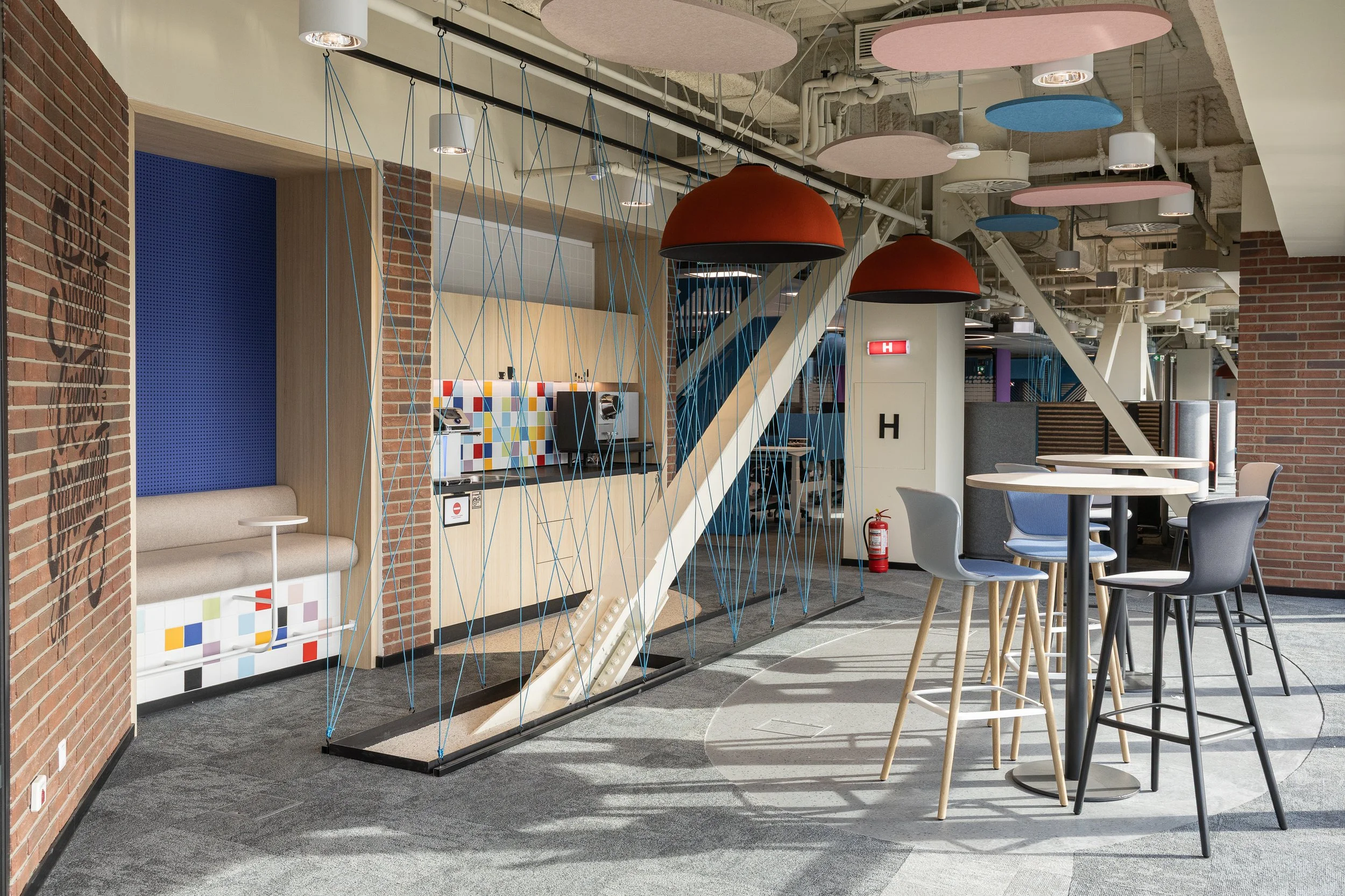 Interior of a modern office or cafe space with brick walls, high tables, and colorful chairs. There are decorative ceiling panels in pink and blue, and a rope climbing structure near a booth with a built-in seat.