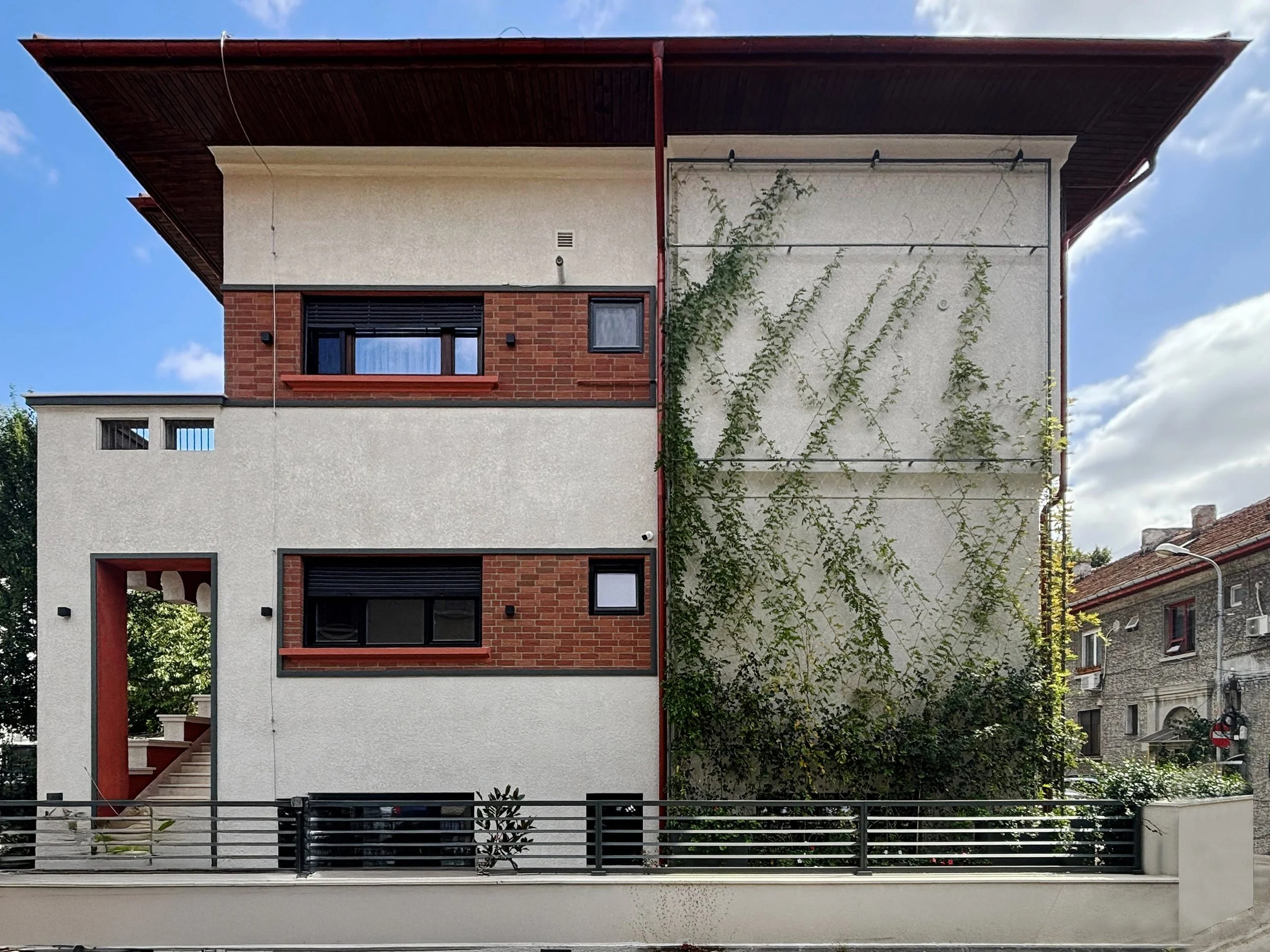 A modern multi-story building with a white facade, red brick accents, and large windows. The right side features a white wall with green climbing vines. The building is surrounded by a black metal fence and has a staircase on the left side. The sky is partly cloudy.